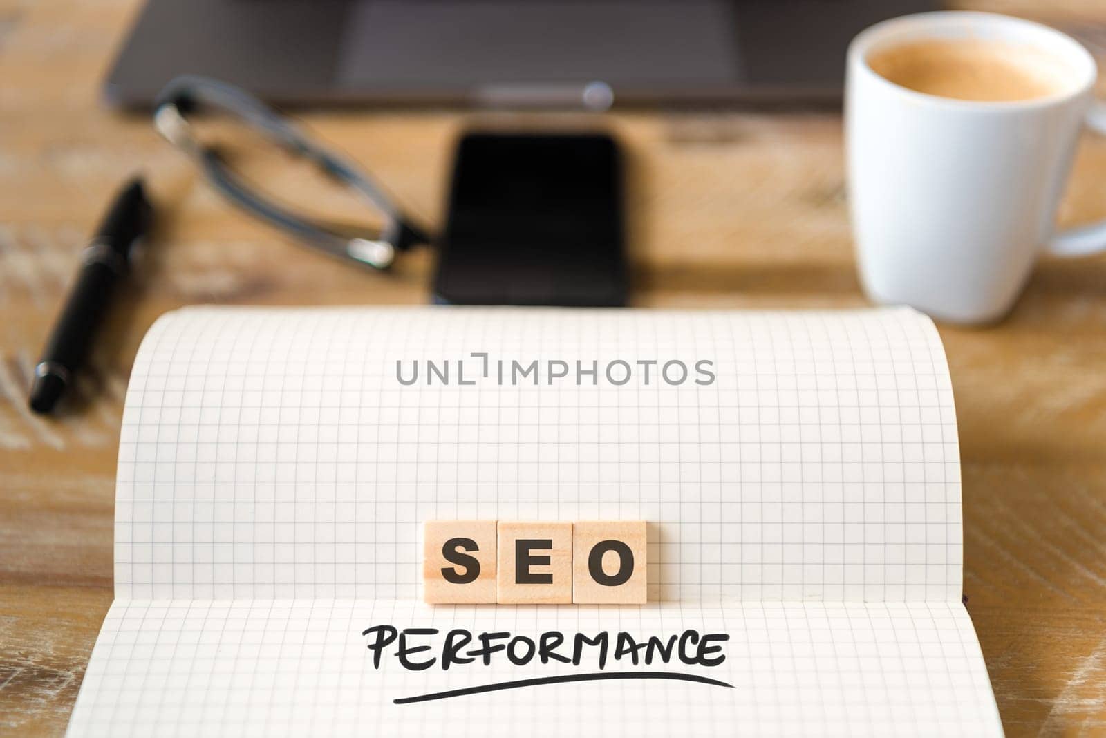 Closeup on notebook over wood table background, focus on wooden blocks with letters making SEO Performance text. Business concept image. Laptop, glasses, pen and mobile phone in defocused background