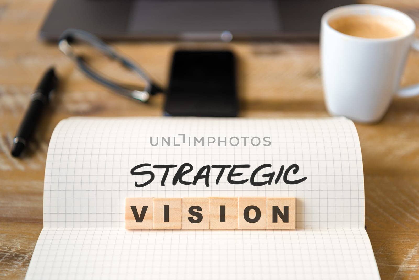 Closeup on notebook over wood table background, focus on wooden blocks with letters making Strategic Vision text. Business concept image. Laptop, glasses, pen and mobile phone in defocused background