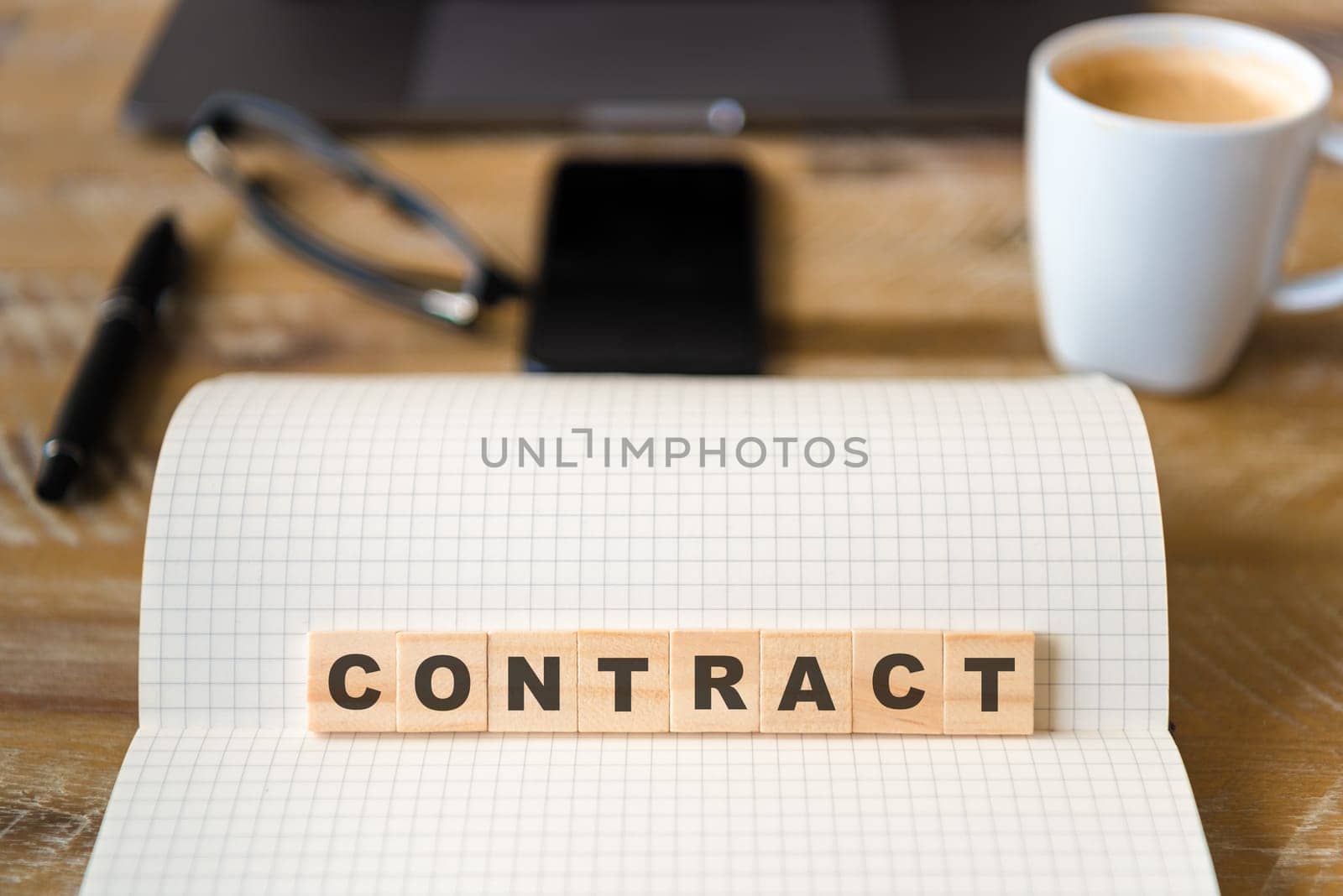 Closeup on notebook over wood table background, focus on wooden blocks with letters making CONTRACT word. Business concept image. Laptop, glasses, pen and mobile phone in defocused background