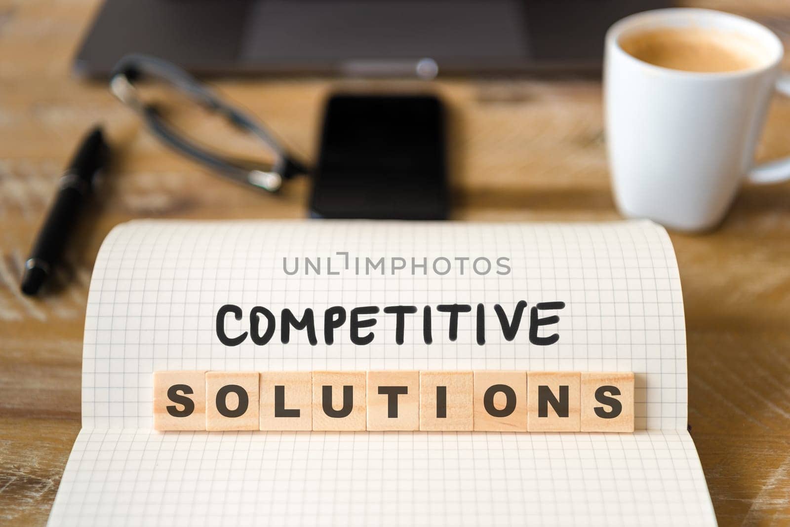 Closeup on notebook over wood table background, focus on wooden blocks with letters making Competitive Solutions text. Business concept image. Laptop, glasses, pen and mobile phone in defocused background