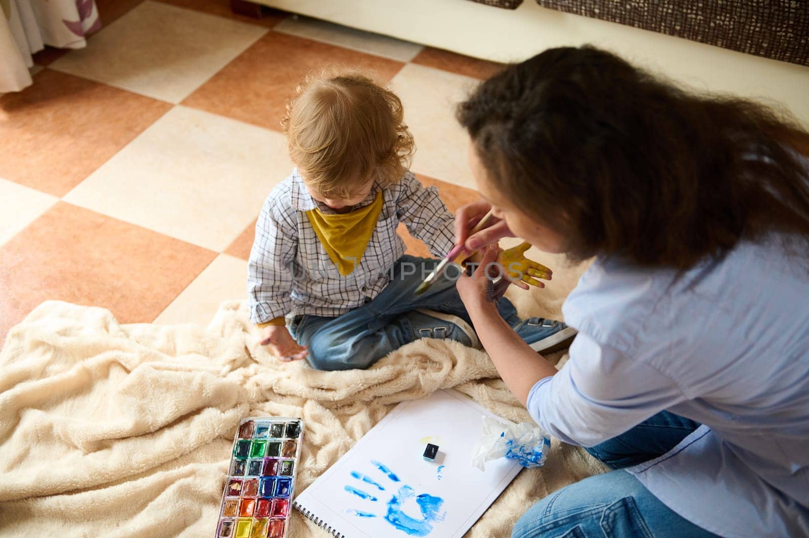 A mother engages with her child in a creative painting activity, using watercolors to paint on paper and their hands, depicting joyful moments and parental bonding.