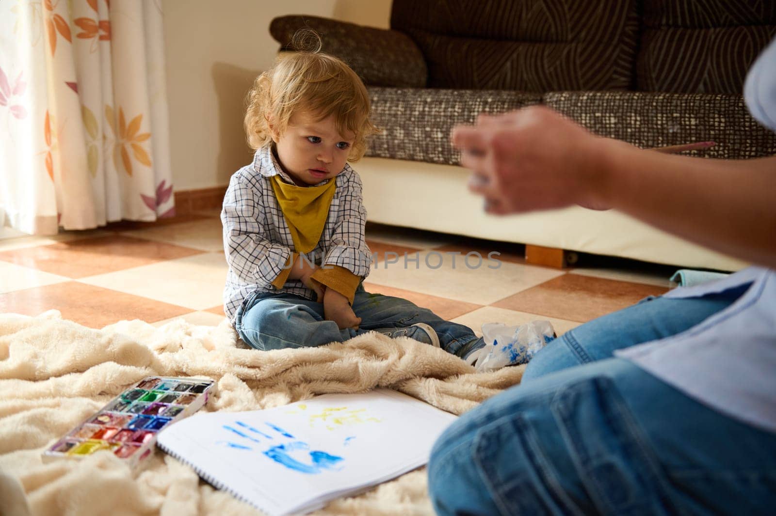 A young child on a blanket paints on paper, exploring creativity and learning under parental guidance in a comfortable home environment.