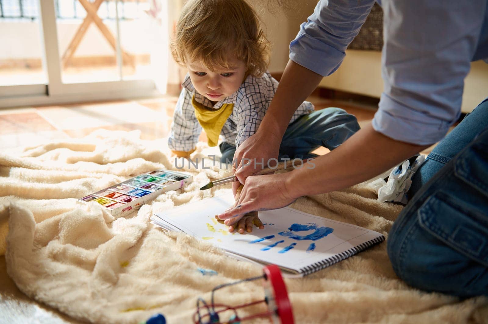 A mother and her young child enjoy creative bonding time, painting vibrant handprints on paper while sitting together in a comfortable and warm home space.