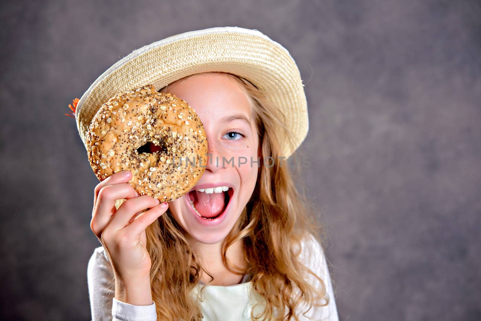 nice blond girl with straw hat in front of gray background looking through a big bagel