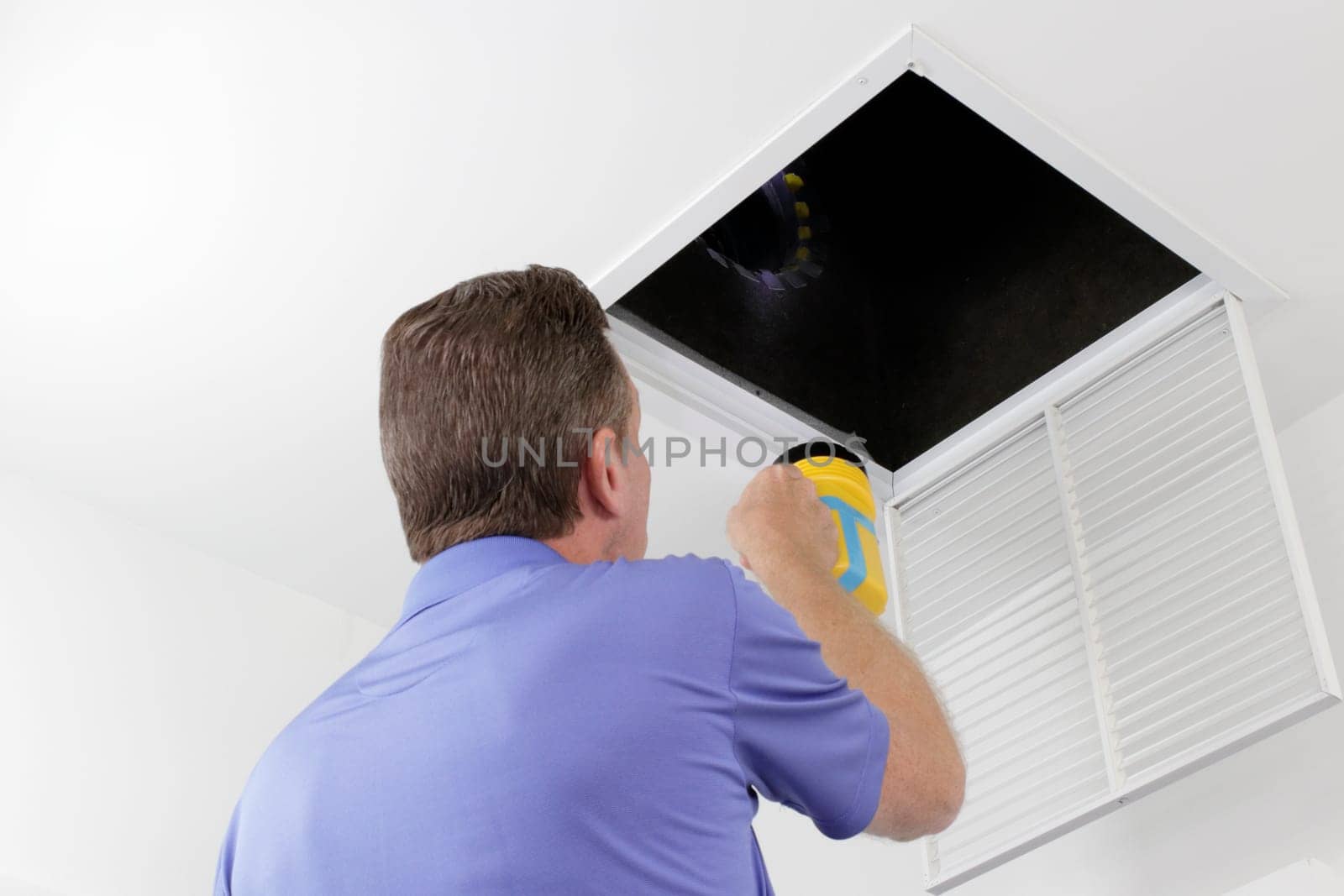 Older male with a yellow flashlight examining HVAC ducts in a large square vent. Male technician looking over the air ducts inside a home air intake vent.