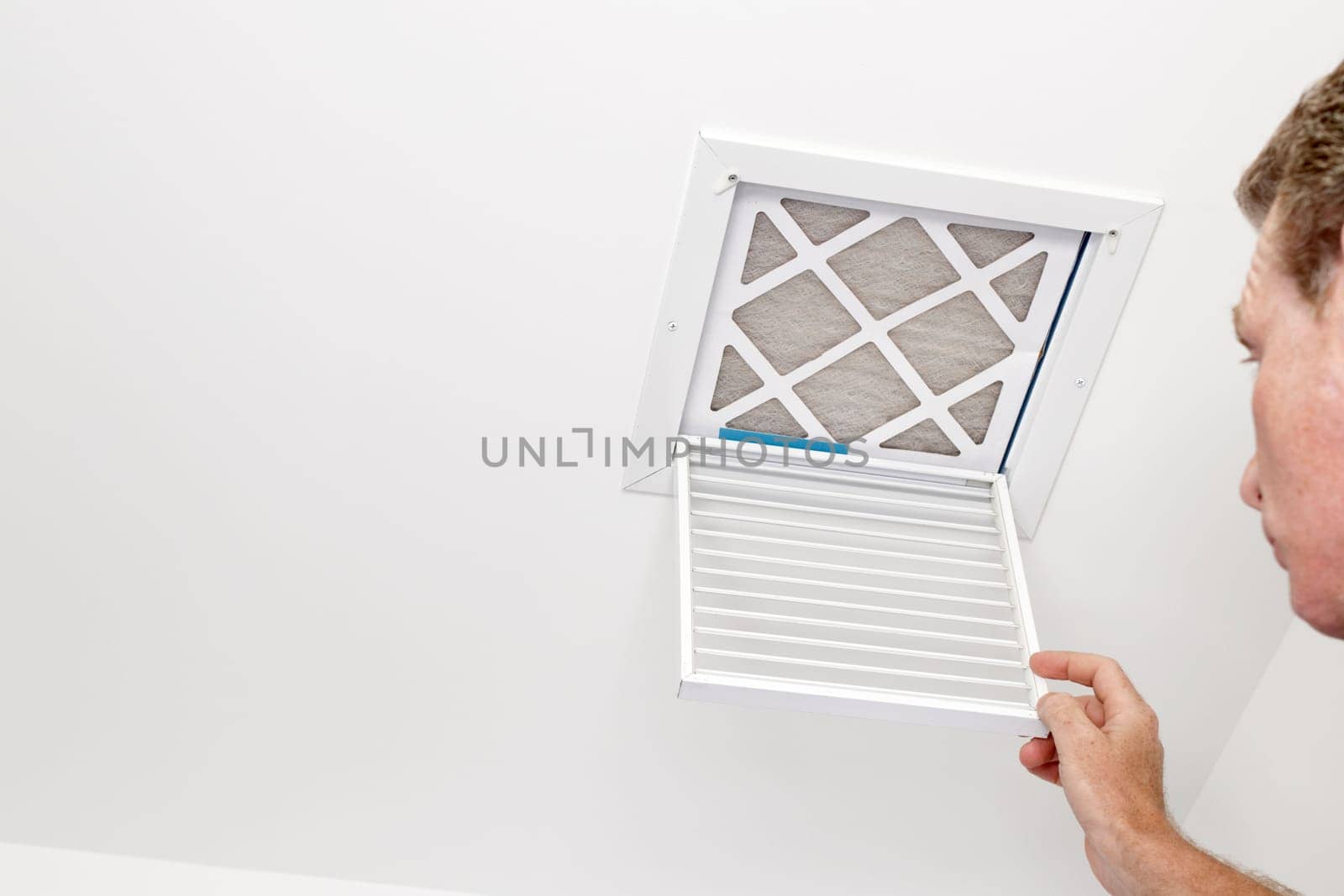Mature adult opening a small square HVAC air intake vent revealing a dirty air filter in the ceiling. Man opened a a vent grill cover to a dirty ceiling air filter..