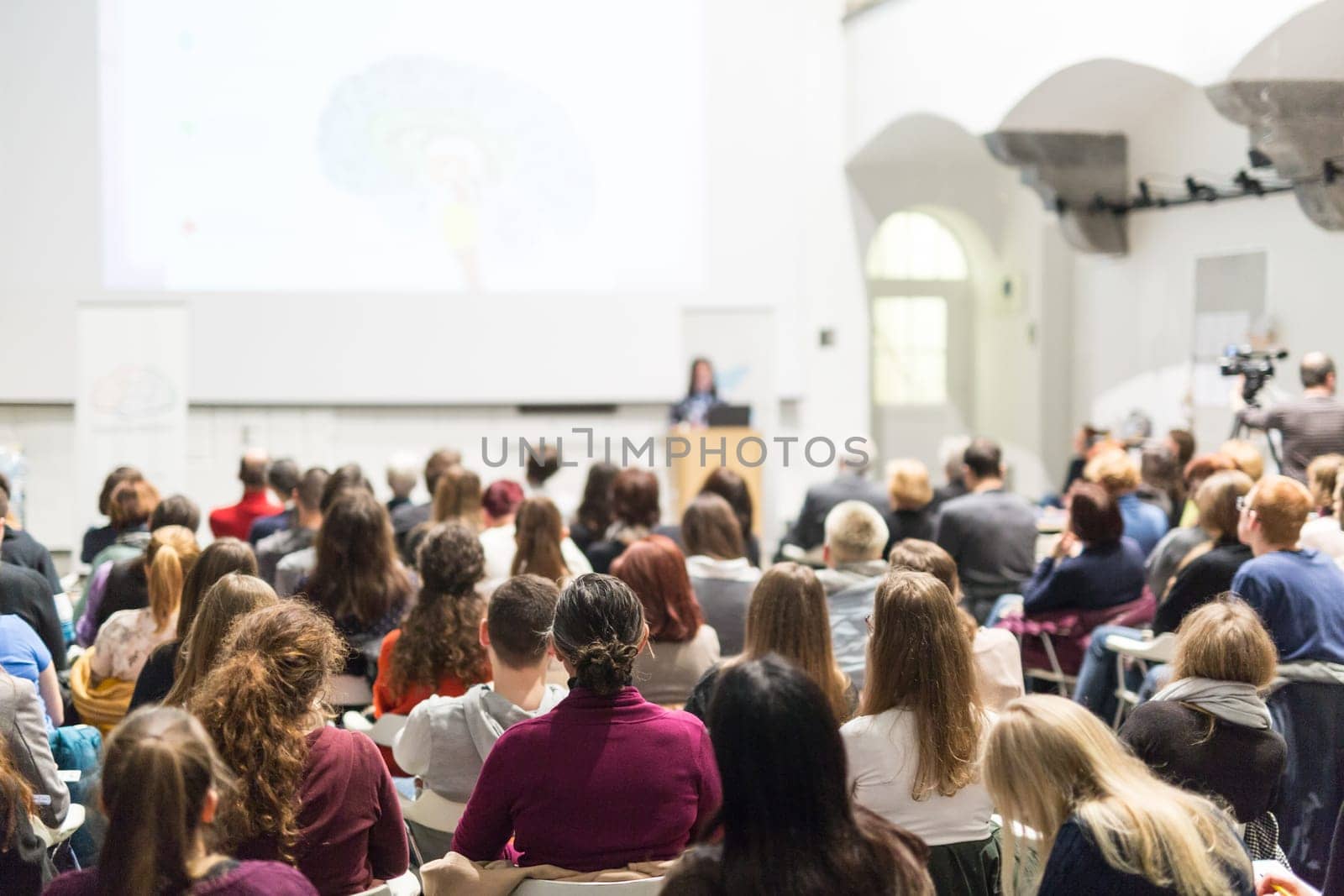 Female speaker giving presentation in lecture hall at university workshop. Audience in conference hall. Rear view of unrecognized participant in audience. Scientific conference event.