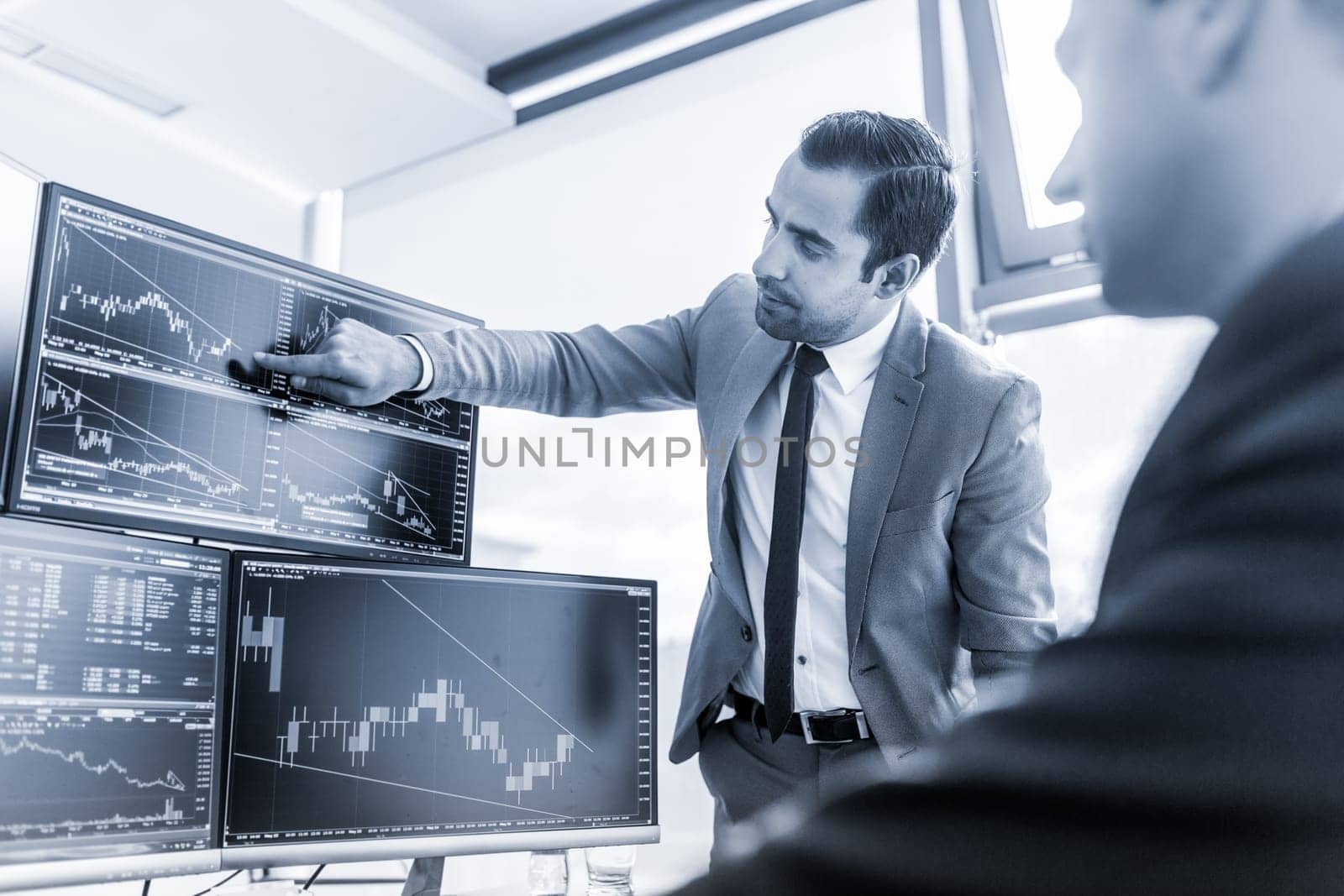 Businessmen trading stocks online. Stock brokers looking at graphs, indexes and numbers on multiple computer screens. Colleagues in discussion in traders office. Blue toned black and white.