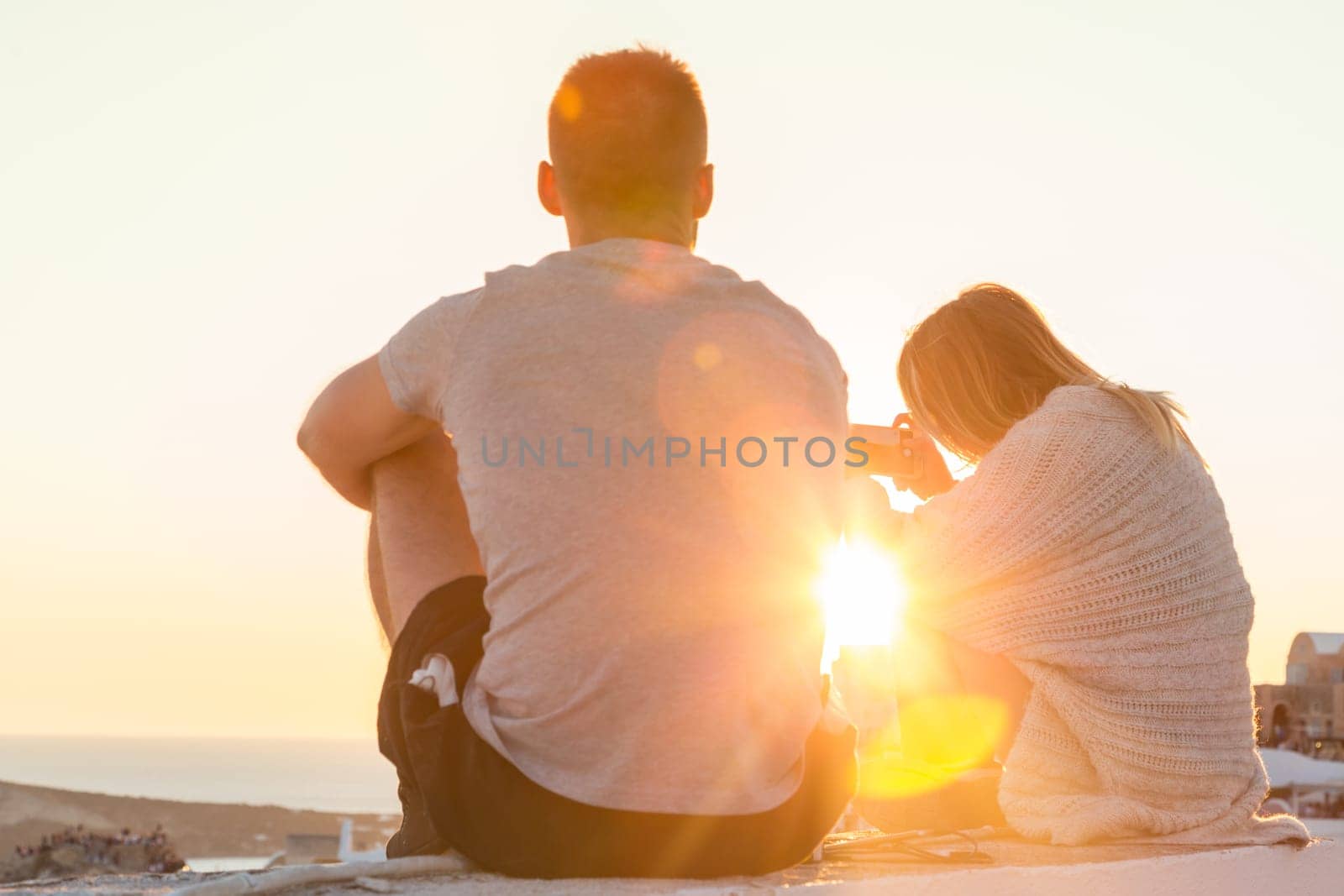 Rear view of unrecognizable young couple watching sunrise and taking vacation photos at Santorini island, Greece.