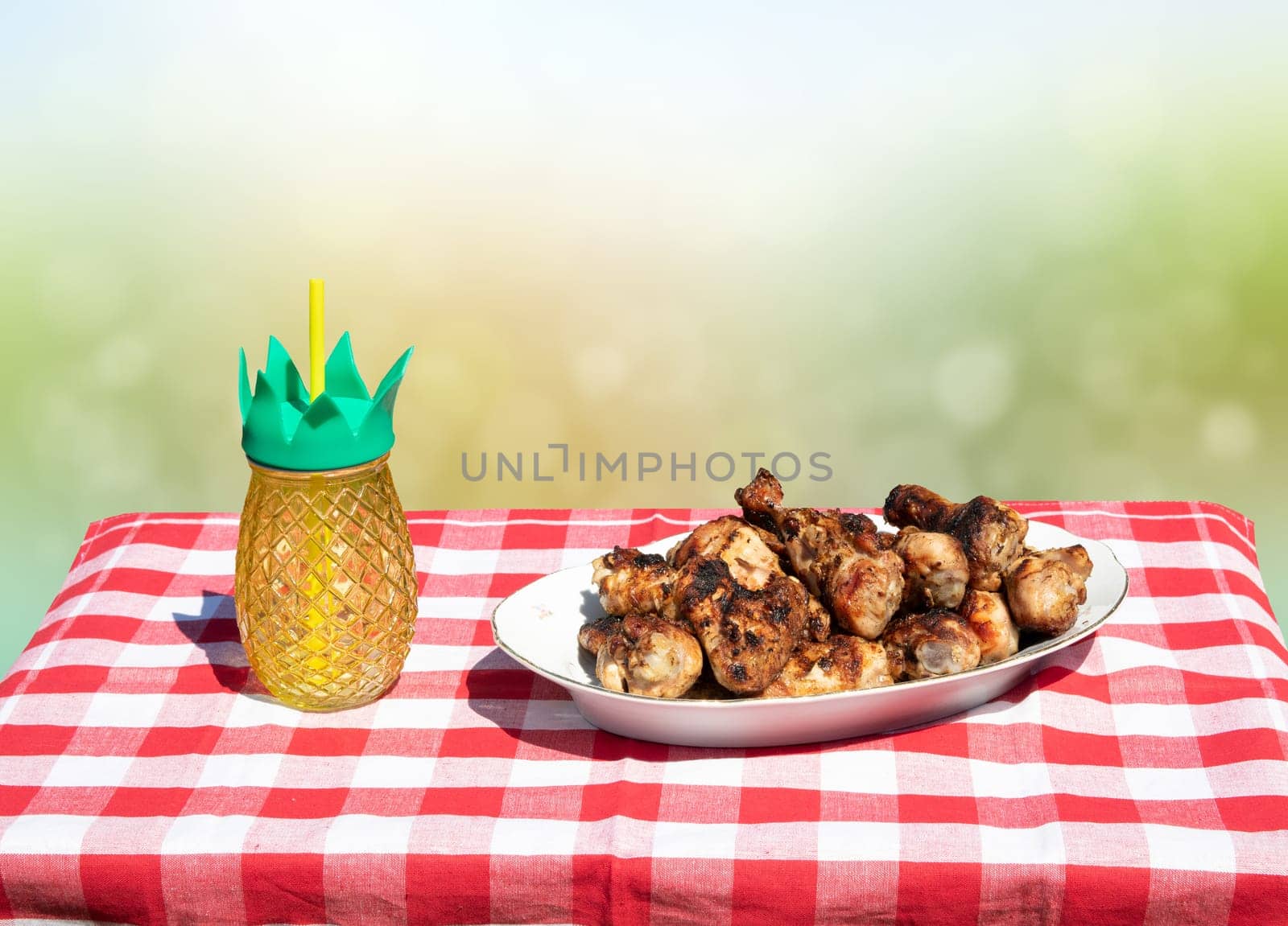 Fresh grilled chicken meat and a glass of juice on table with red checkered tablecloth and bright green white bokeh background. BBQ.