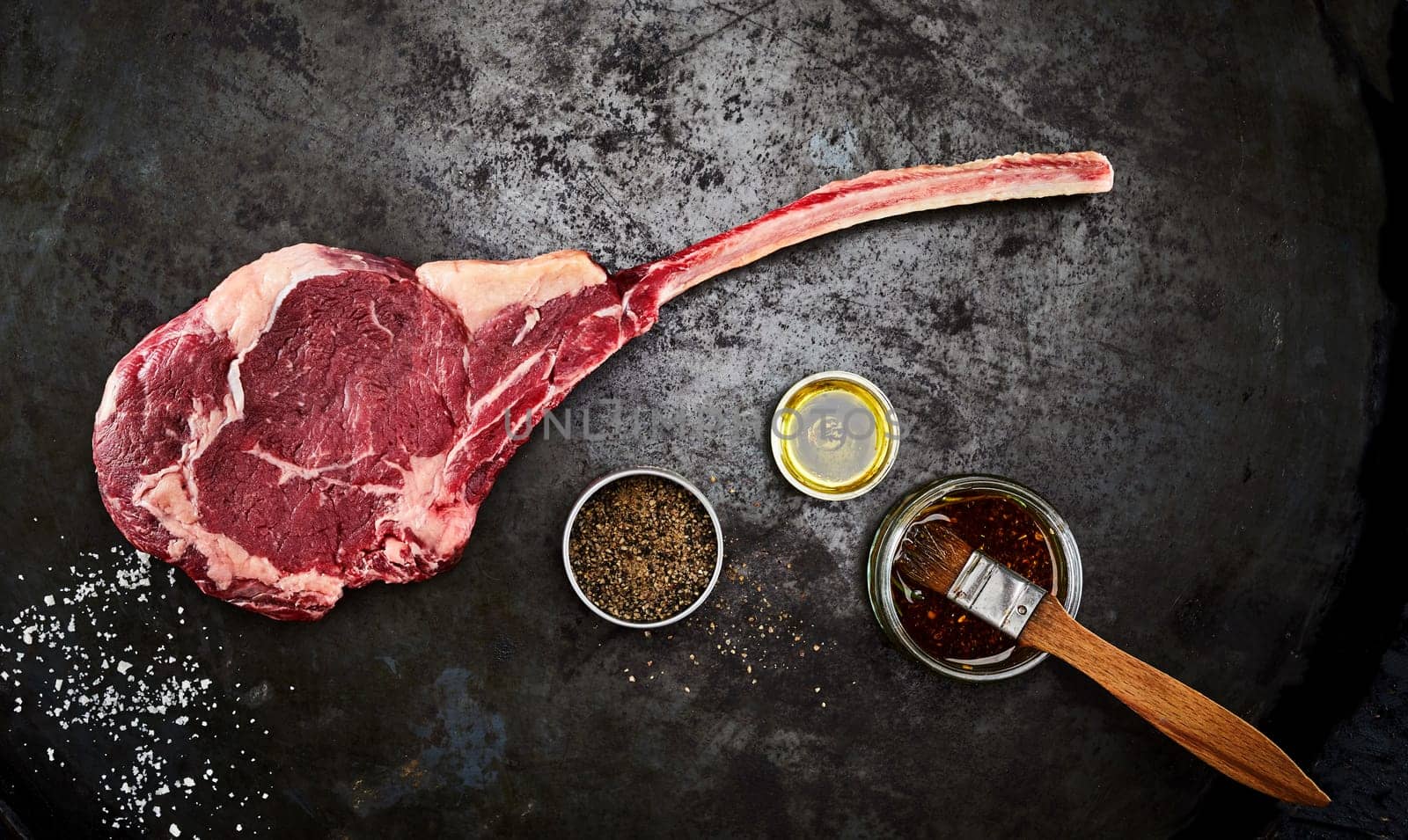 Tender raw trimmed bone-in ribeye steak with jars of marinade, spice rub an olive oil viewed from above on an old metal surface