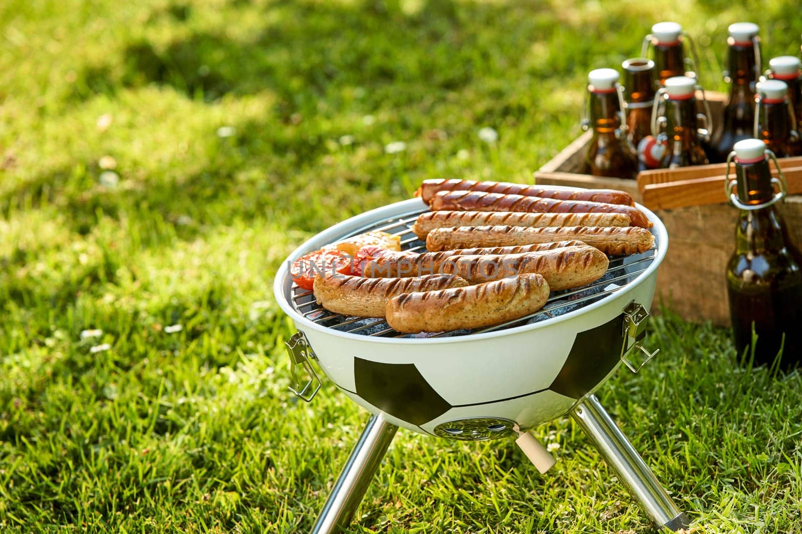 Sausages grilling on a portable BBQ shaped like a black and white soccer ball alongside a crate of beers outdoors in a field for a World Cup supporter celebration