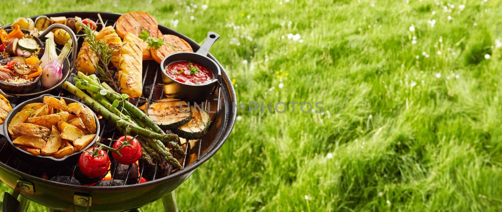 Assortment of fresh healthy vegetables on a BBQ grilling over a hot fire in a green grassy spring or summer field in banner format with copy space