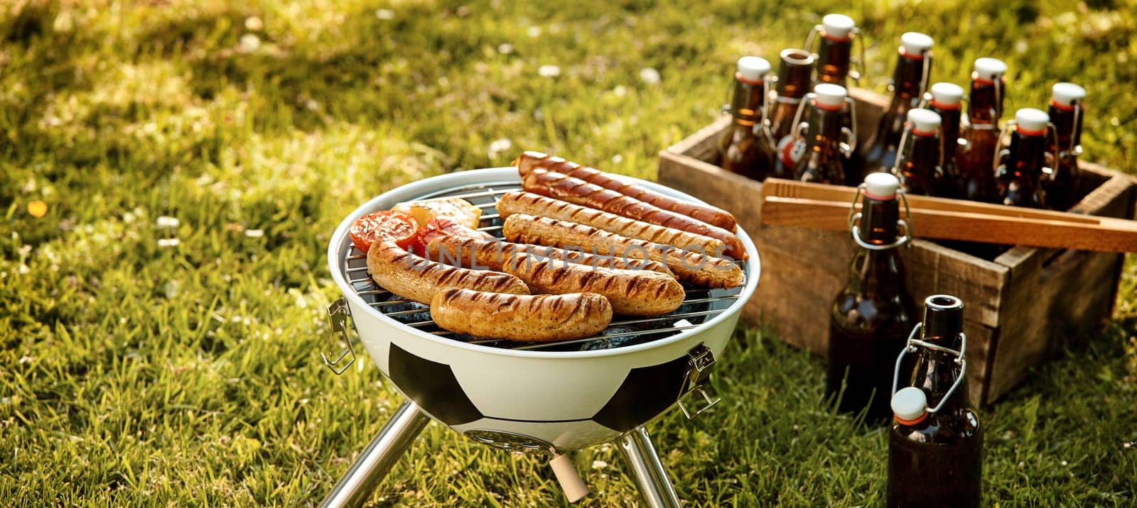 Soccer World Cup themed barbecue with pork and beef sausages grilling over a fire in a soccer ball BBQ and crate of beers in a green field in panorama format