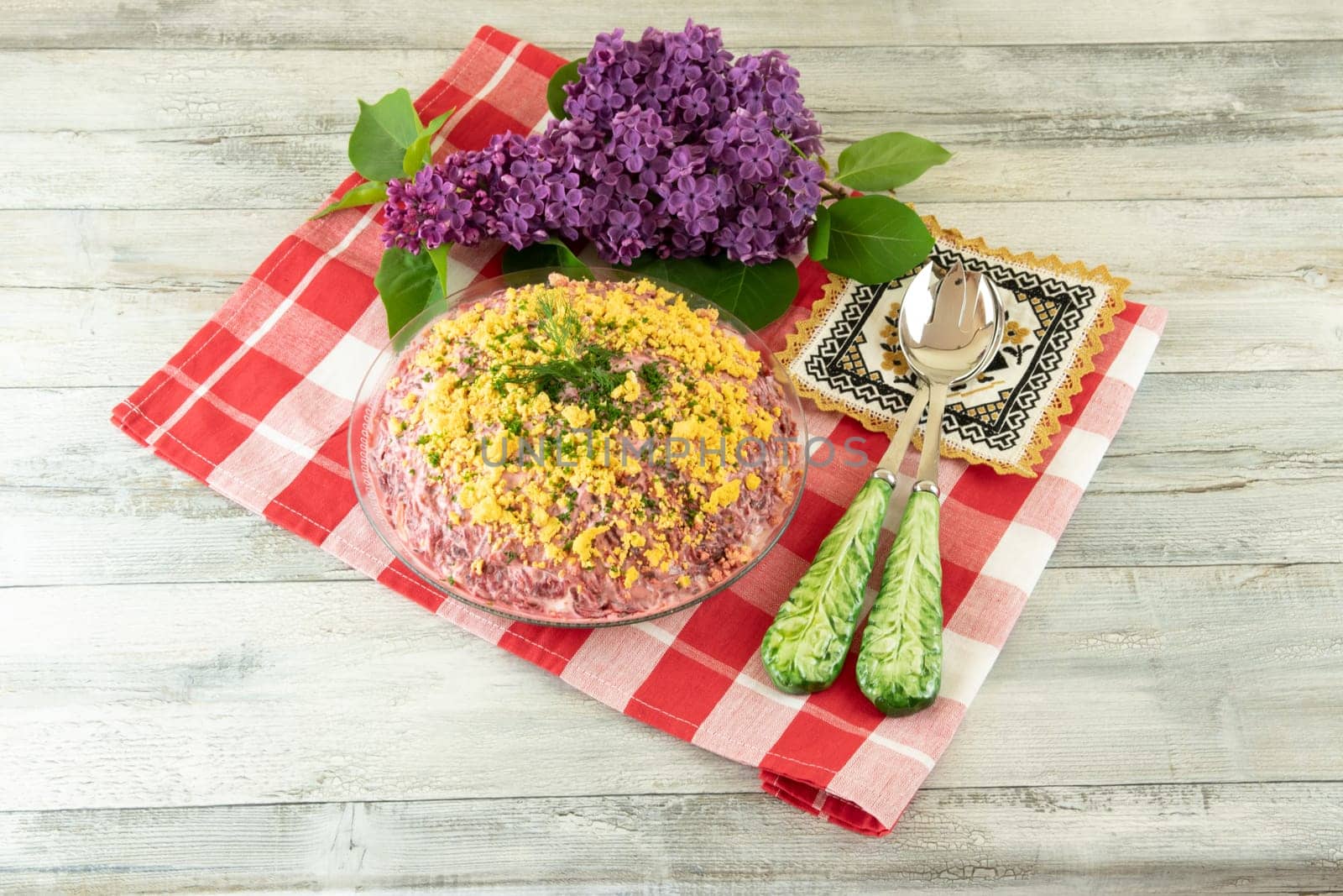 Traditional russian layered salad is named herring under a fur coat. Traditional Russian cuisine presented at red checkered tablecloth and decorated with a fresh purple lilac branch and two spoons. Concept Russian food.