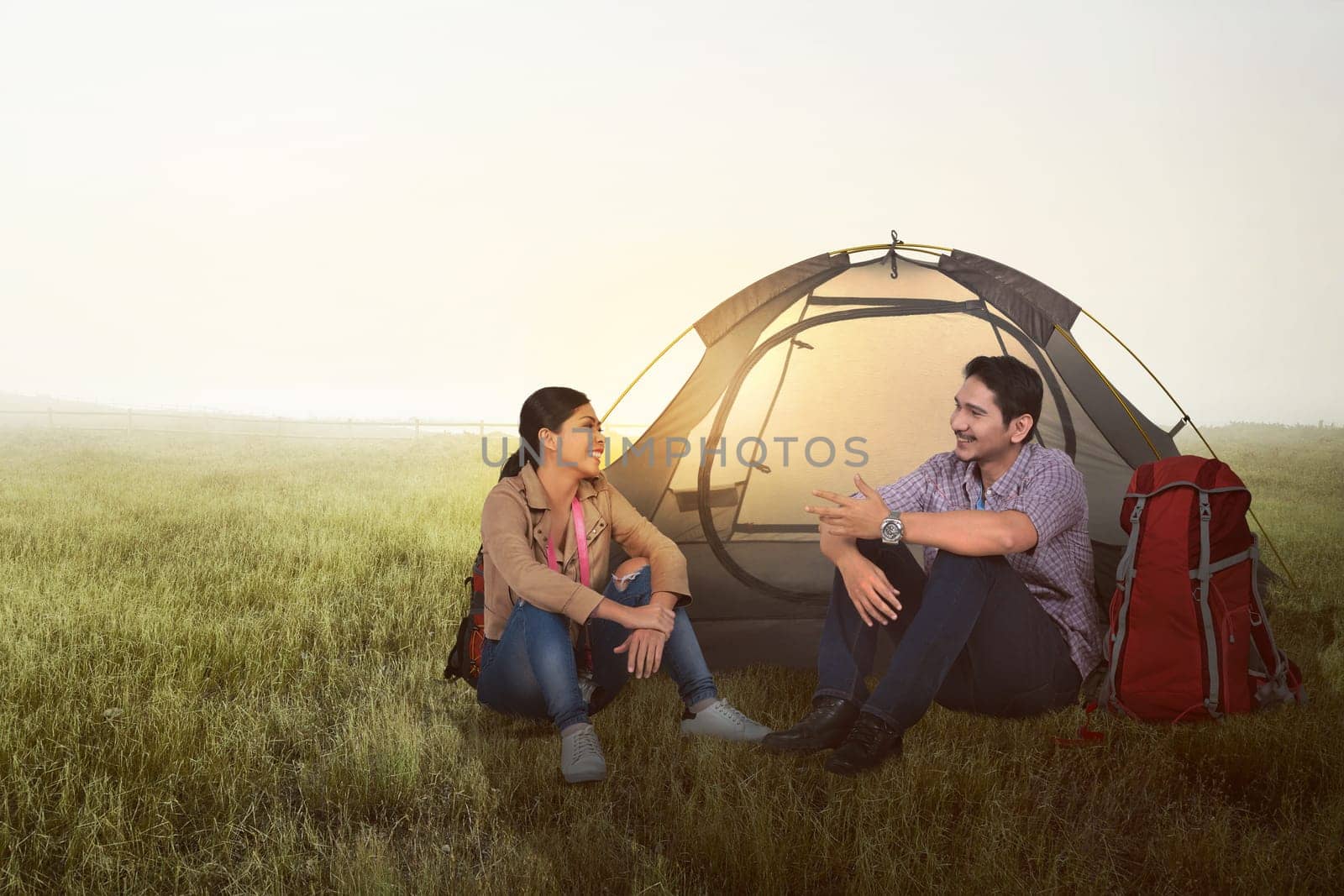 Smiling asian couple camping with backpack and tent on the meadow field