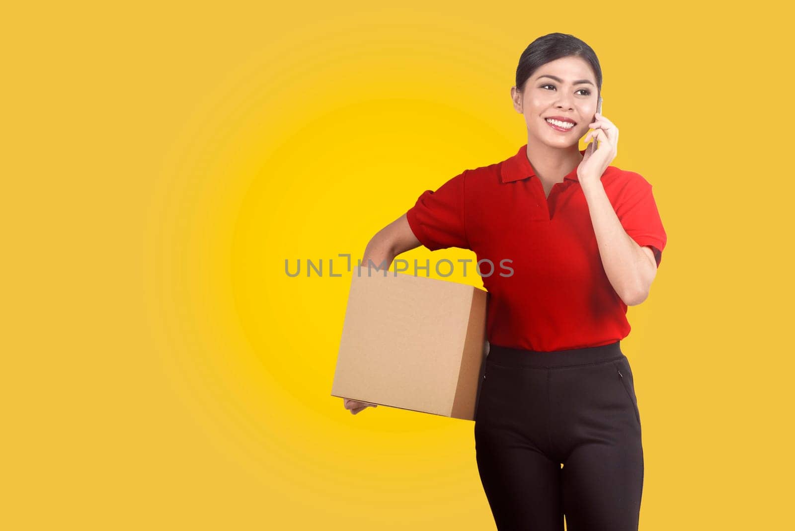 Pretty asian female courier using mobile phone while holding parcel over yellow background