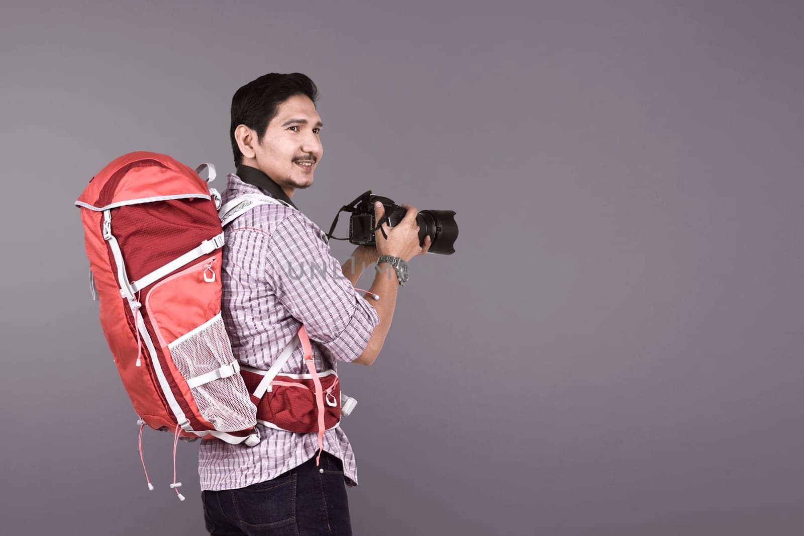 Young asian photographer with backpack holding camera over gray background