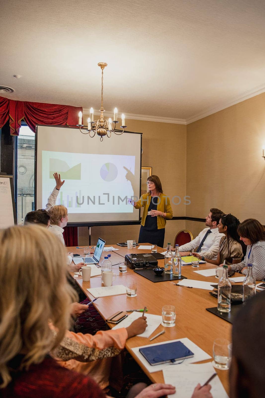 Rear view of a conference room filled with business people. One businessman is raising his hand to ask the presenter a question.