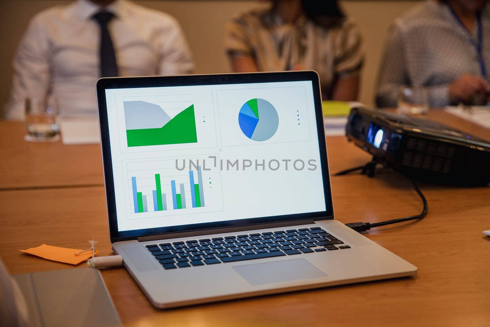 Front view of a computer plugged into a projector showing charts and graphs with information on. Unrecognisable people in the background.