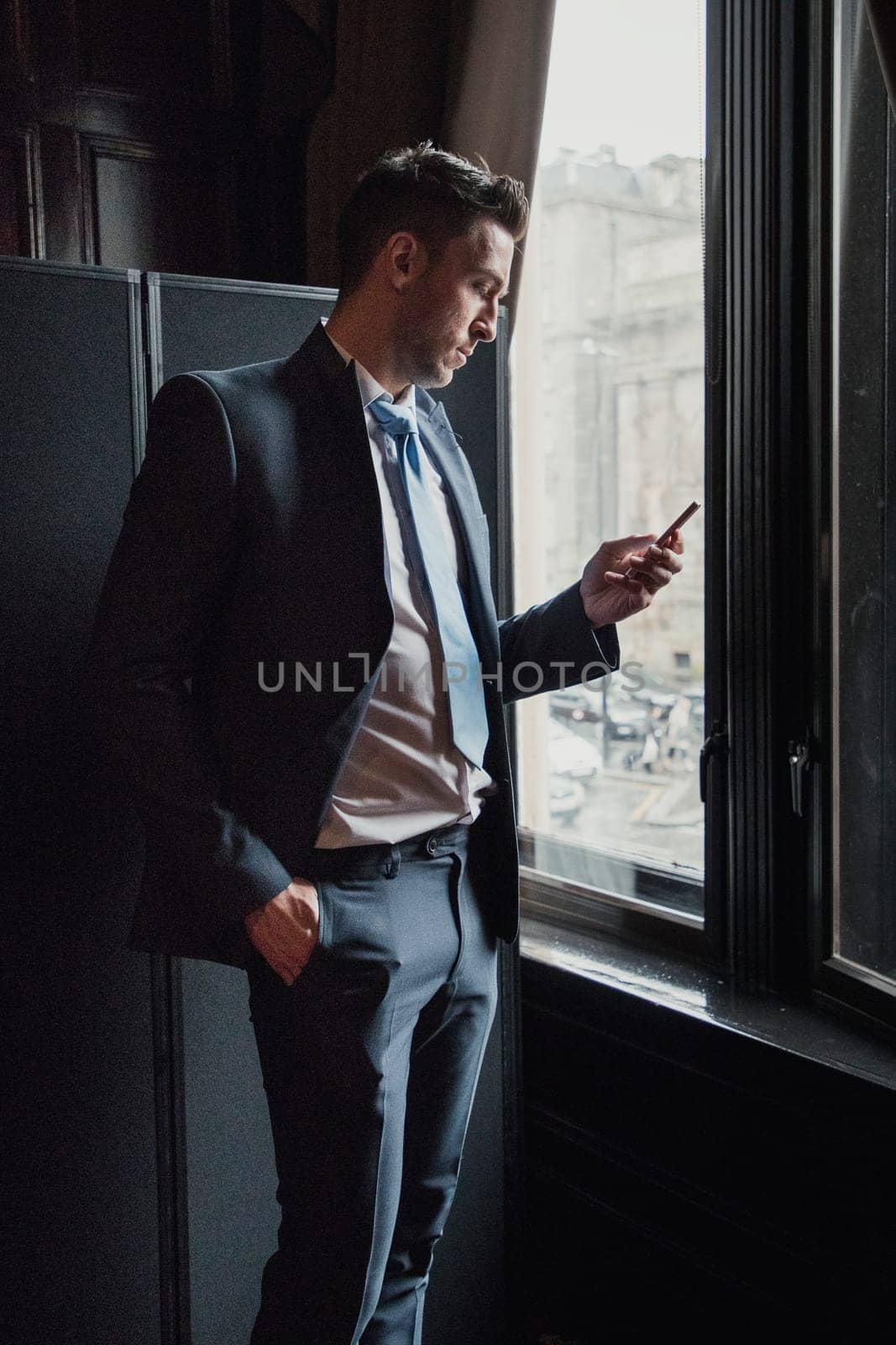 Side view of a young businessman standing next to a window in the office. He is checking his smart phone while waiting.