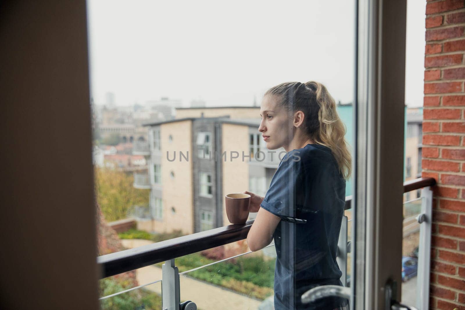 Rear, through the window view of a young female adult in her casual clothing enjoying a cup of tea on the balcony.
