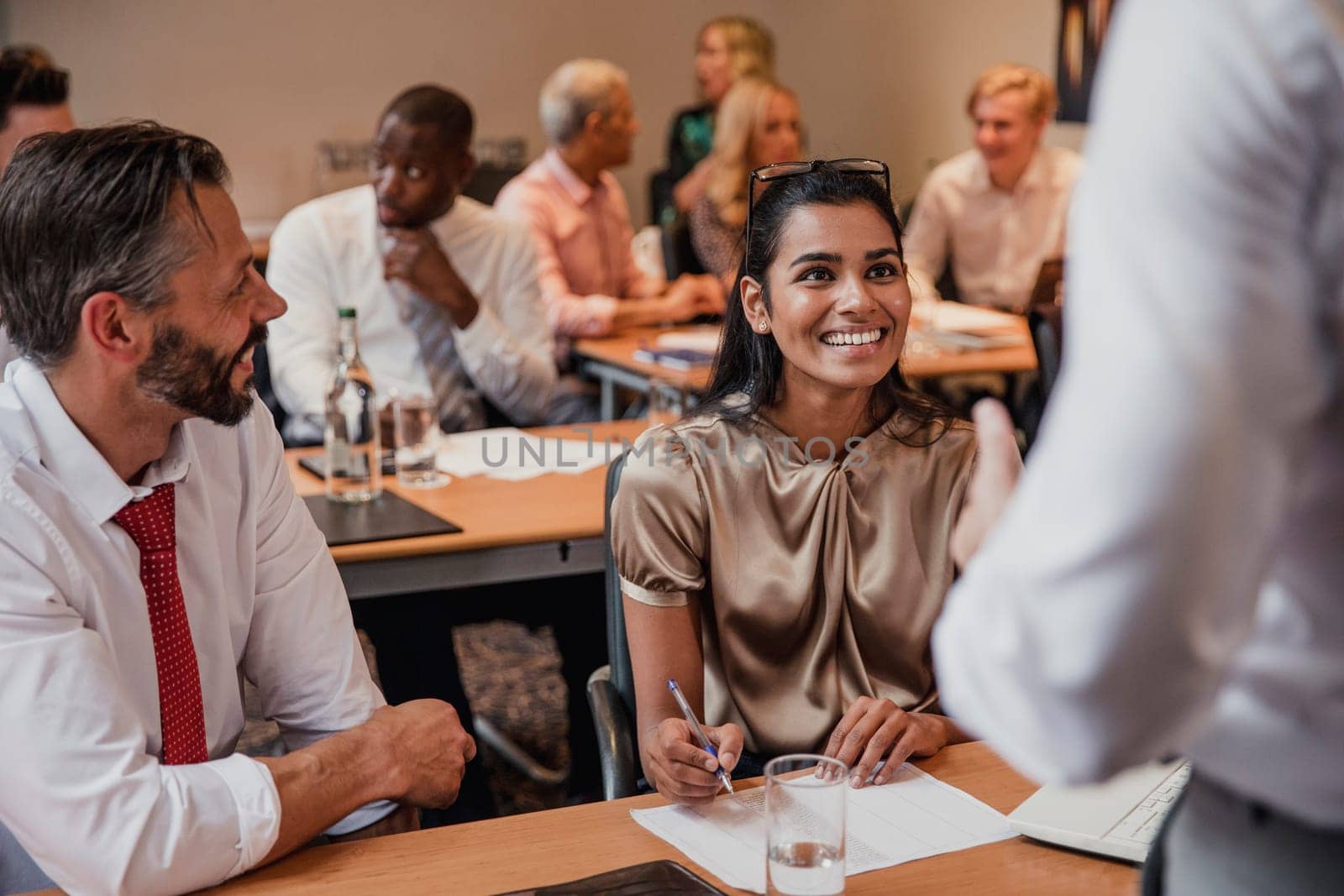 Front view of a businessman and businesswoman sitting at a table in a business meeting. An unrecognisable person stands in the right hand of the frame.