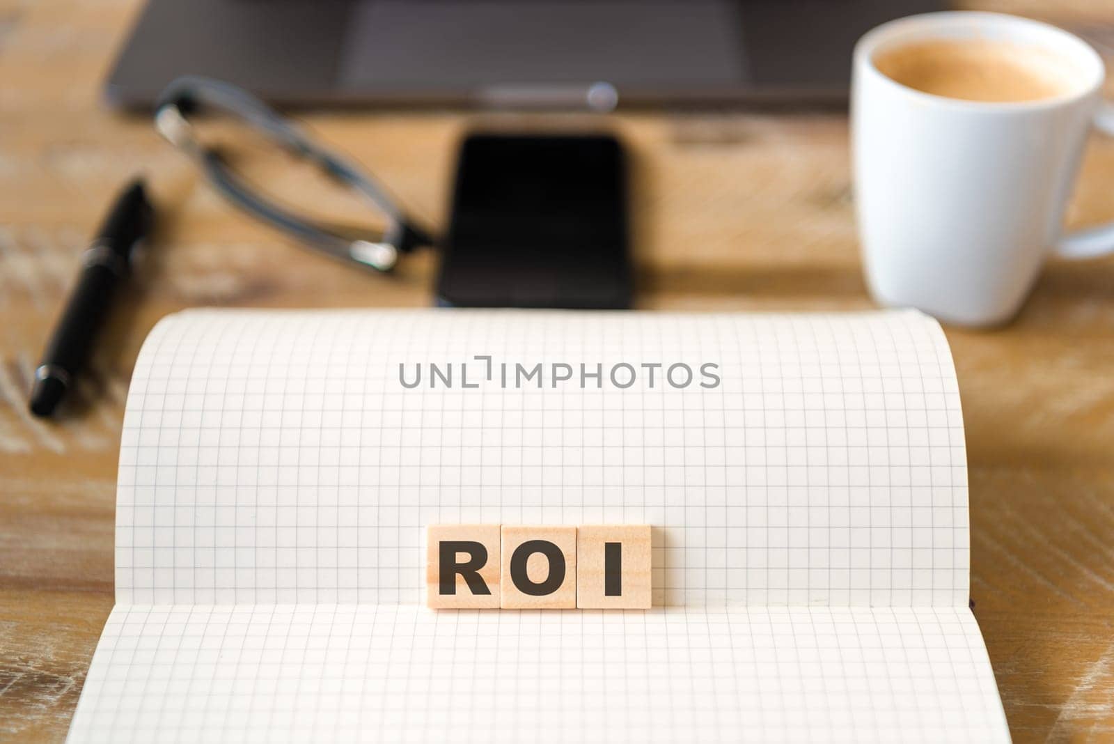 Closeup on notebook over wood table background, focus on wooden blocks with letters making ROI text. Concept image. Laptop, glasses, pen and mobile phone in defocused background