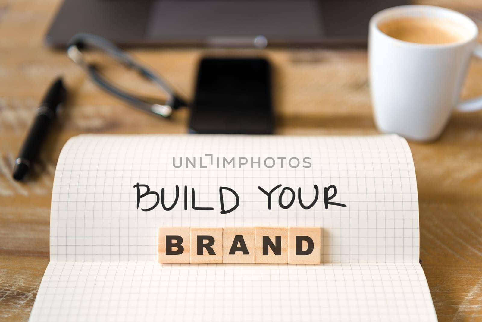 Closeup on notebook over vintage desk surface, front focus on wooden blocks with letters making Build Your Brand text. Business concept image with office tools and coffee cup in background