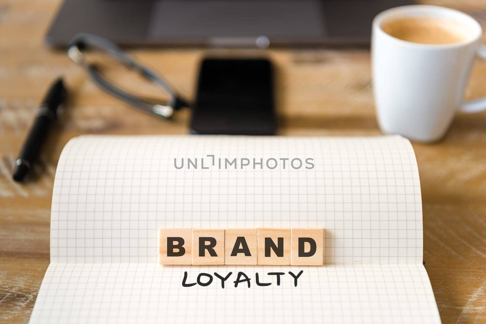 Closeup on notebook over vintage desk surface, front focus on wooden blocks with letters making Brand Loyalty text. Business concept image with office tools and coffee cup in background