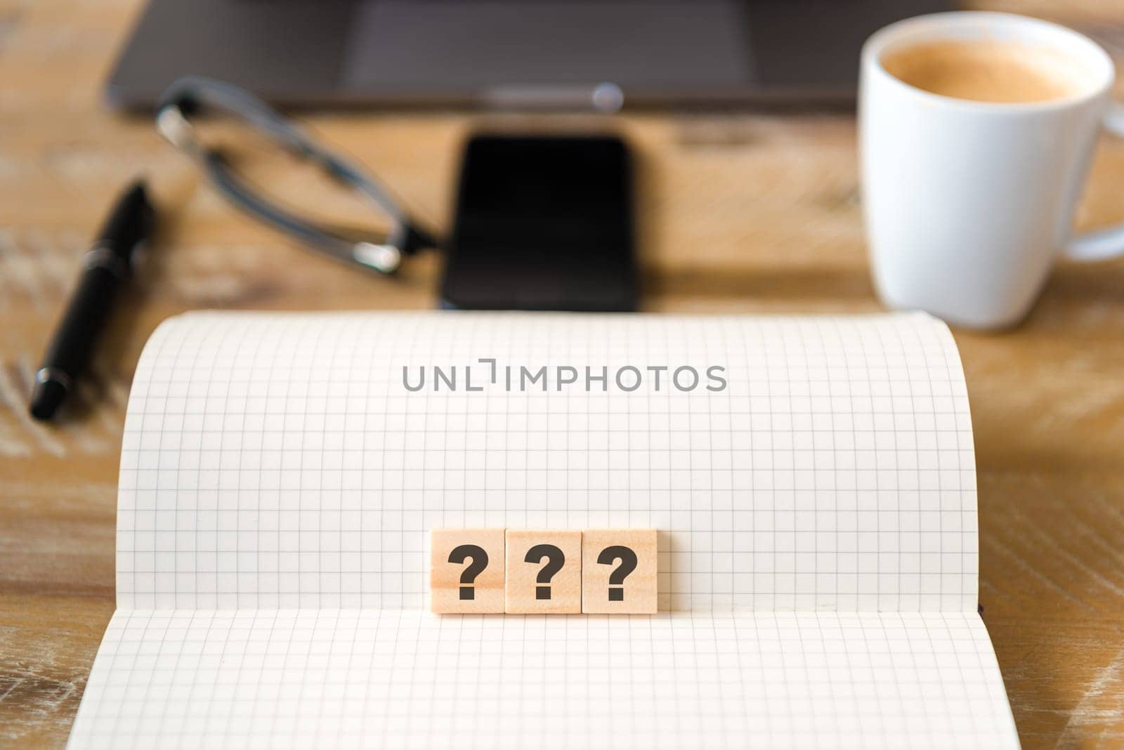 Closeup on notebook over vintage desk surface, front focus on wooden blocks with letters making Three Question Marks text. Business concept image with office tools and coffee cup in background
