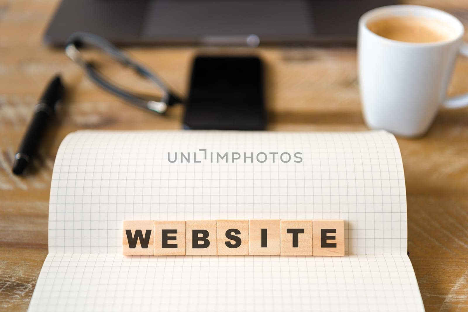 Closeup on notebook over vintage desk surface, front focus on wooden blocks with letters making Website text. Business concept image with office tools and coffee cup in background