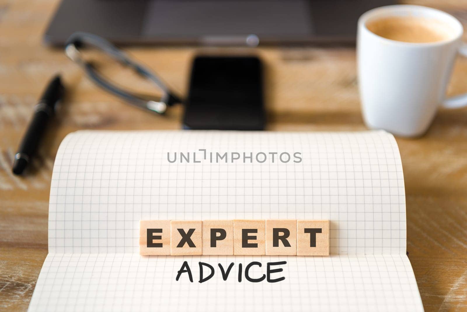 Closeup on notebook over vintage desk surface, front focus on wooden blocks with letters making Expert Advice text. Business concept image with office tools and coffee cup in background