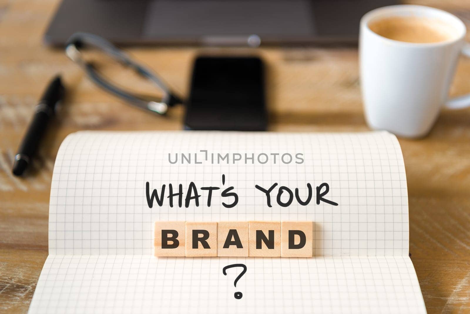 Closeup on notebook over vintage desk surface, front focus on wooden blocks with letters making Whats Your Brand text. Business concept image with office tools and coffee cup in background