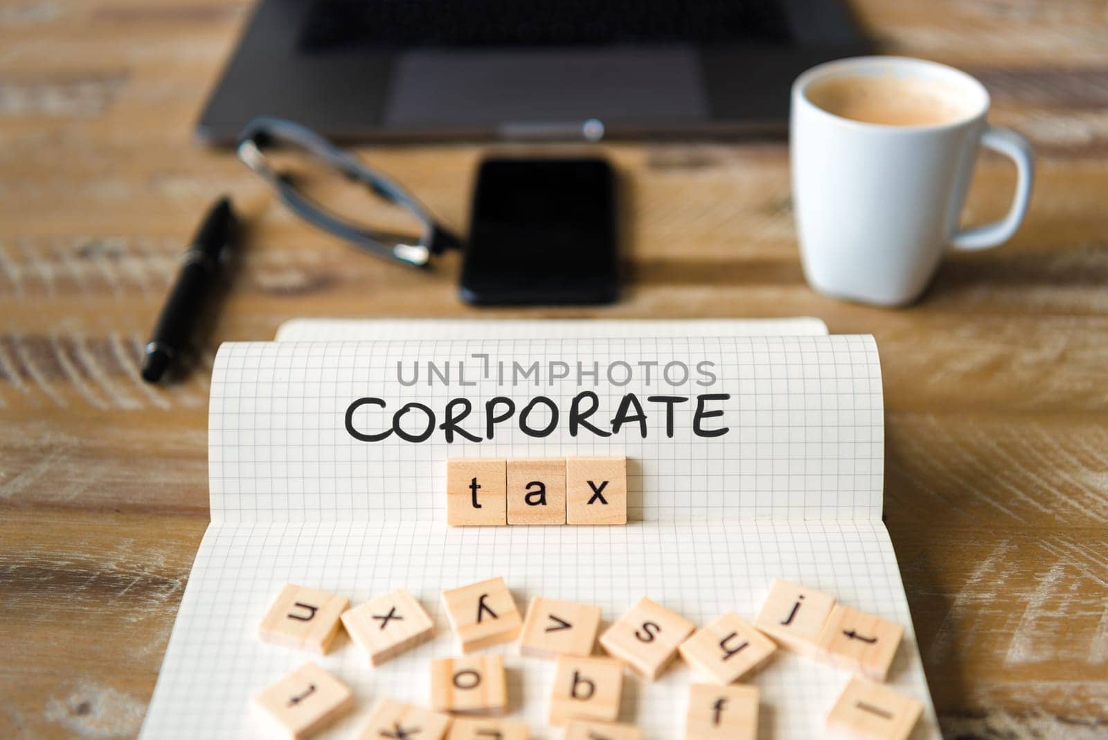 Closeup on notebook over vintage desk surface, front focus on wooden blocks with letters making Corporate Tax text. Business concept image with office tools and coffee cup in background