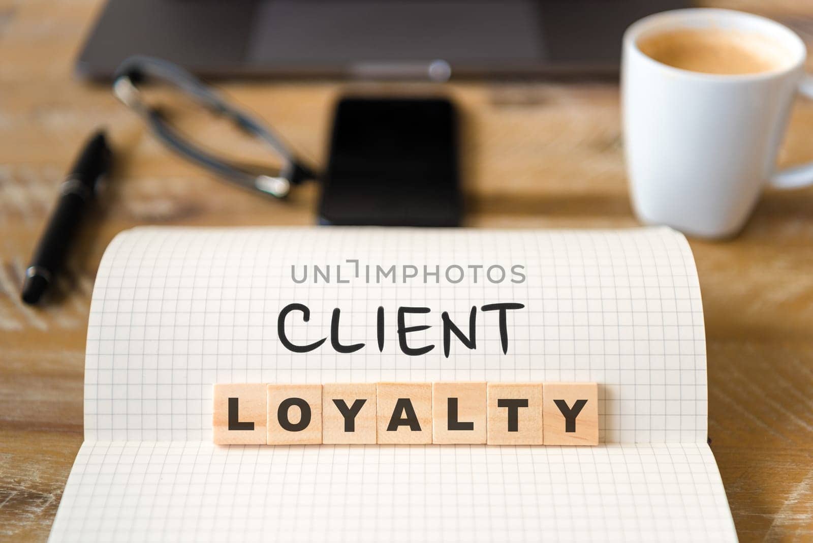 Closeup on notebook over vintage desk surface, front focus on wooden blocks with letters making Client Loyalty text. Business concept image with office tools and coffee cup in background