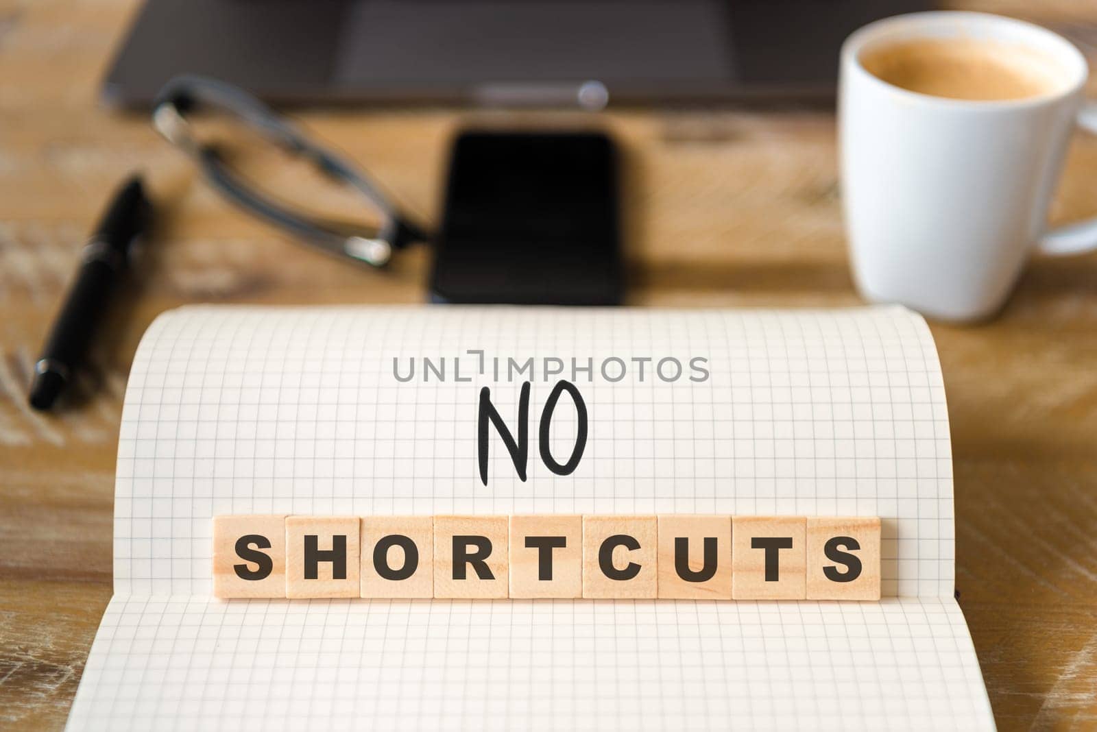 Closeup on notebook over vintage desk surface, front focus on wooden blocks with letters making No Shortcuts text. Business concept image with office tools and coffee cup in background