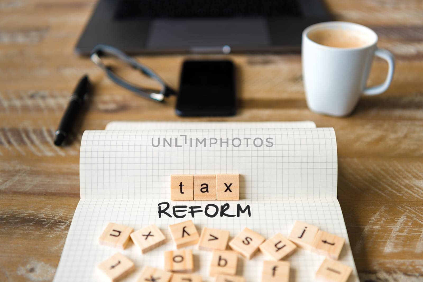 Closeup on notebook over vintage desk surface, front focus on wooden blocks with letters making Tax Reform text. Business concept image with office tools and coffee cup in background