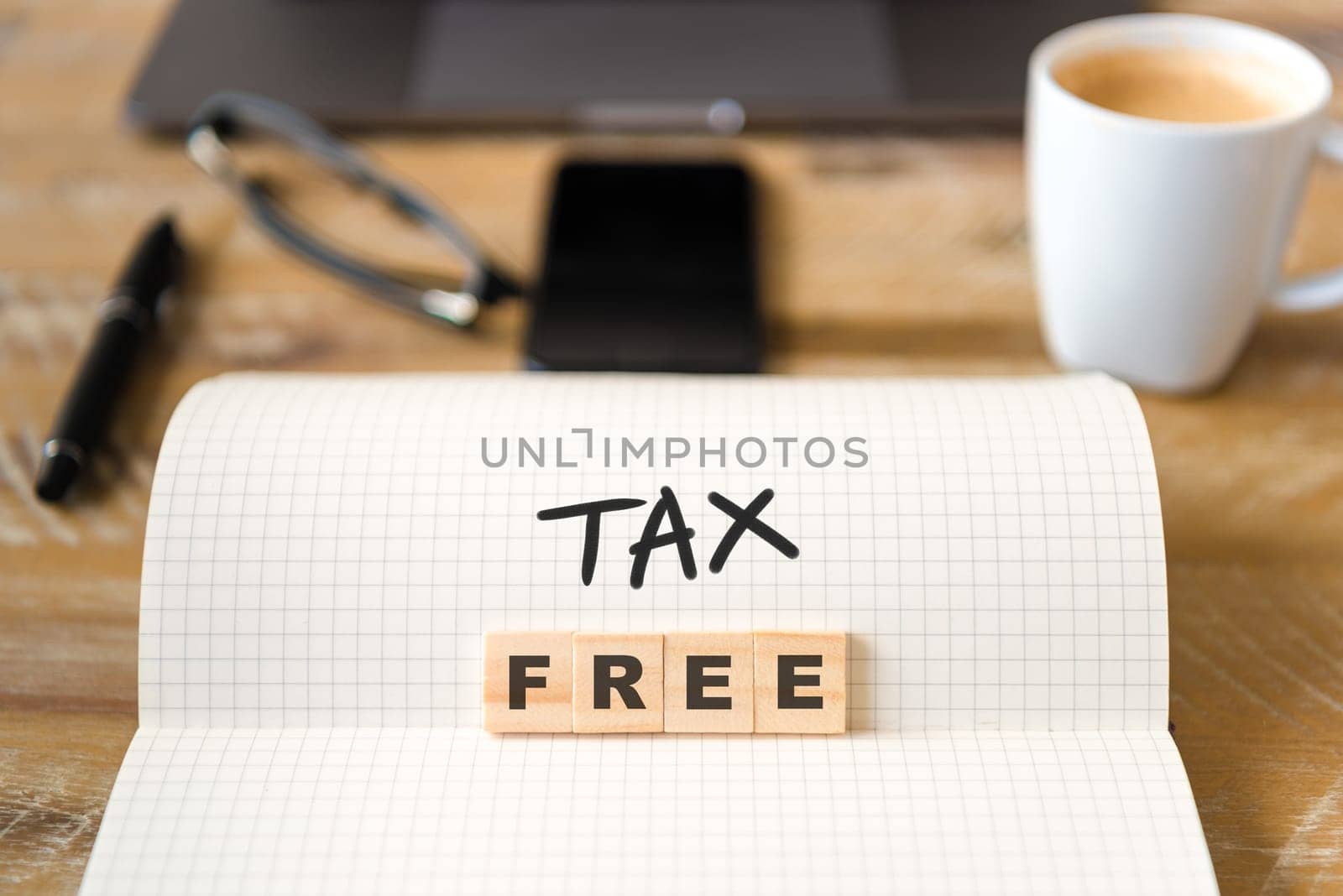 Closeup on notebook over vintage desk surface, front focus on wooden blocks with letters making Tax Free text. Business concept image with office tools and coffee cup in background