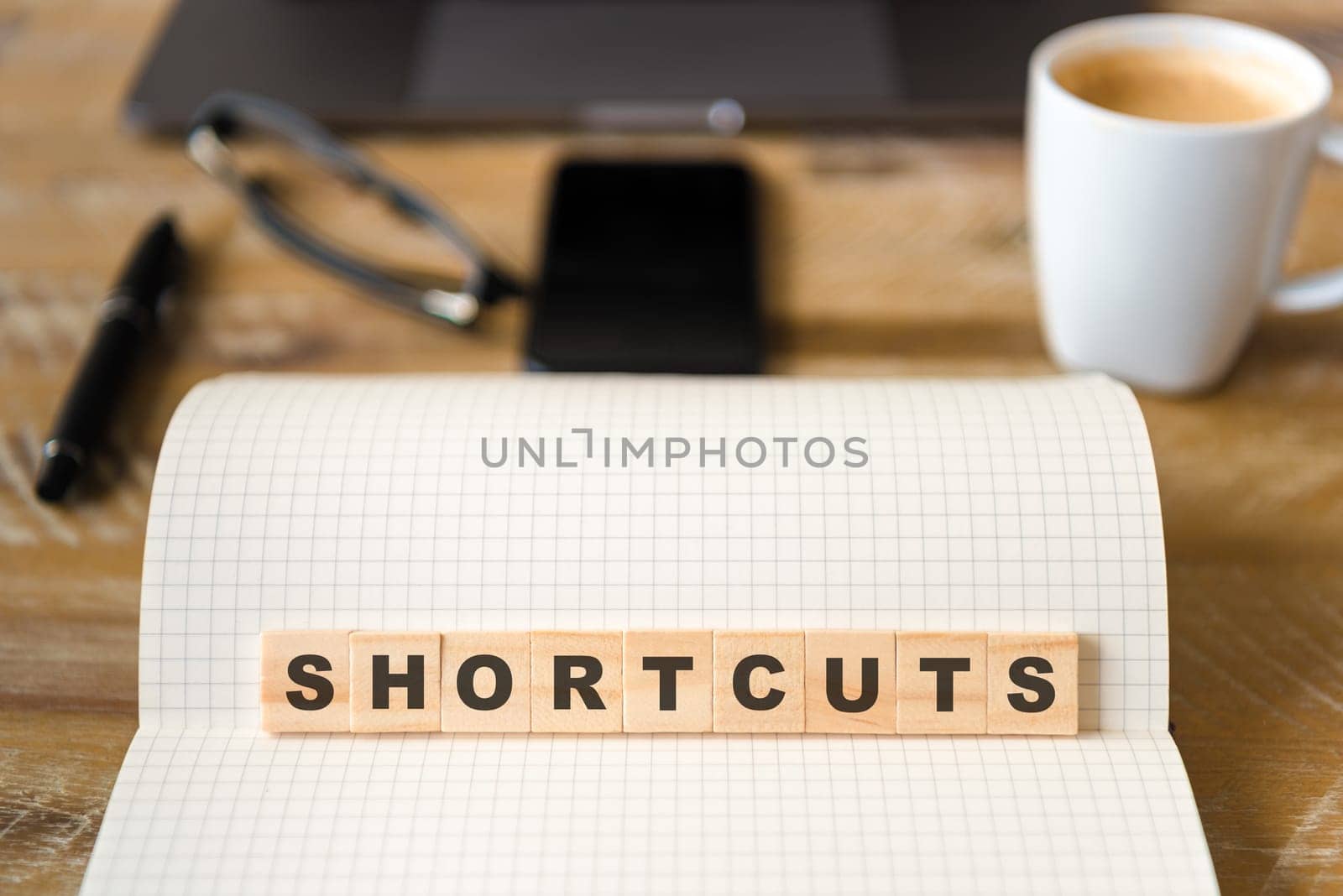 Closeup on notebook over vintage desk surface, front focus on wooden blocks with letters making Shortcuts text. Business concept image with office tools and coffee cup in background