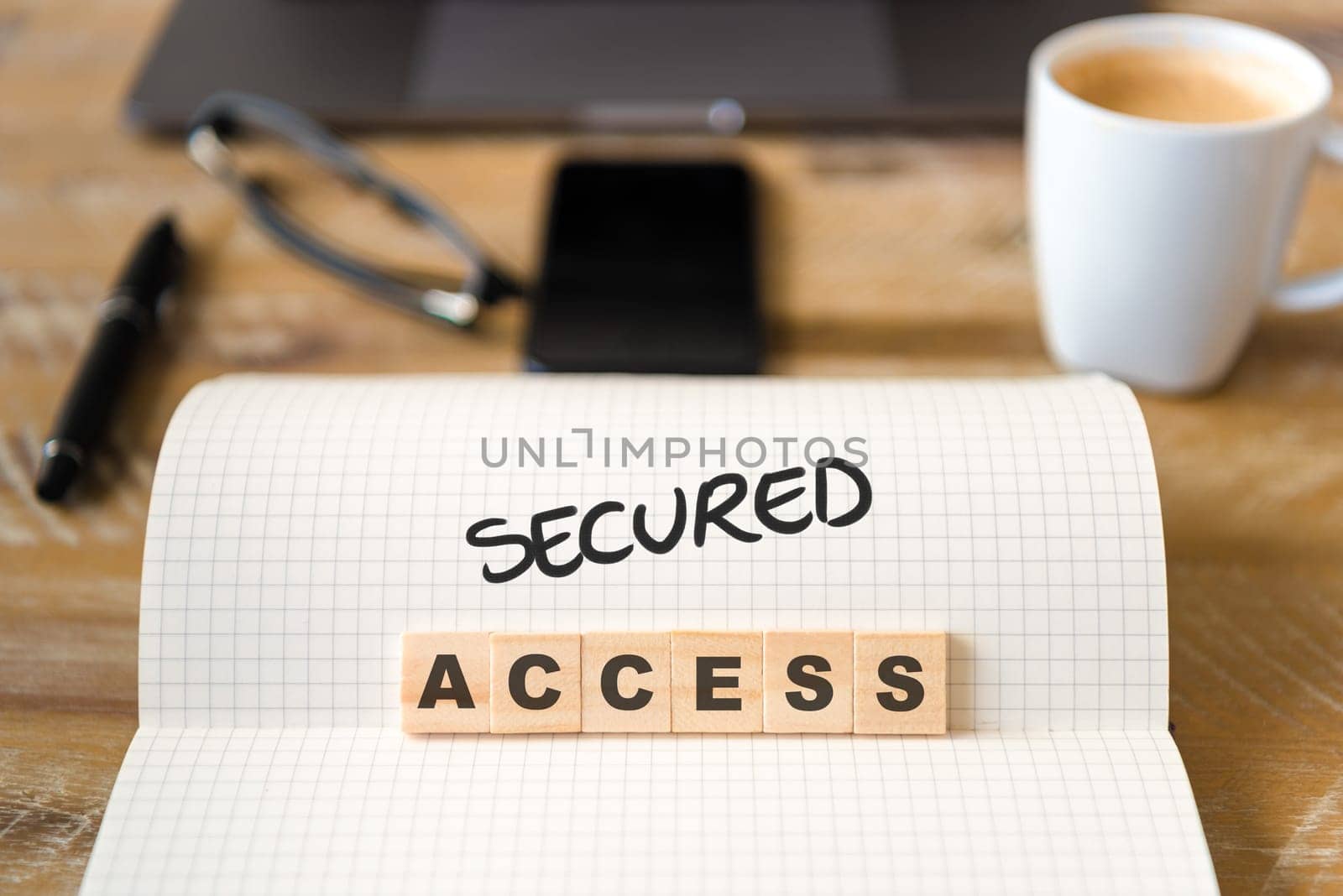 Closeup on notebook over vintage desk surface, front focus on wooden blocks with letters making g Secured Access text. Business concept image with office tools and coffee cup in background
