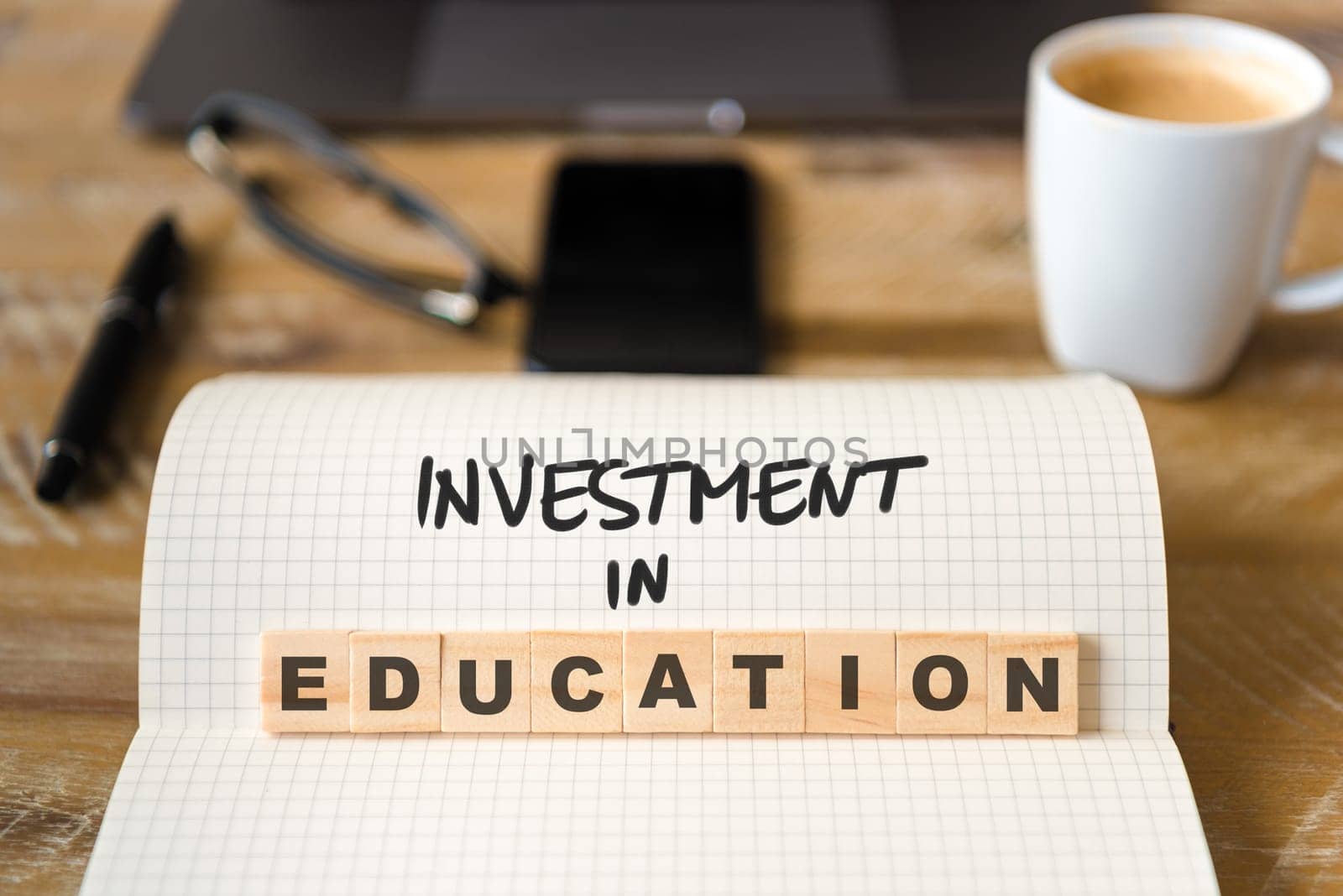 Closeup on notebook over vintage desk surface, front focus on wooden blocks with letters making Investment In Education text. Business concept image with office tools and coffee cup in background