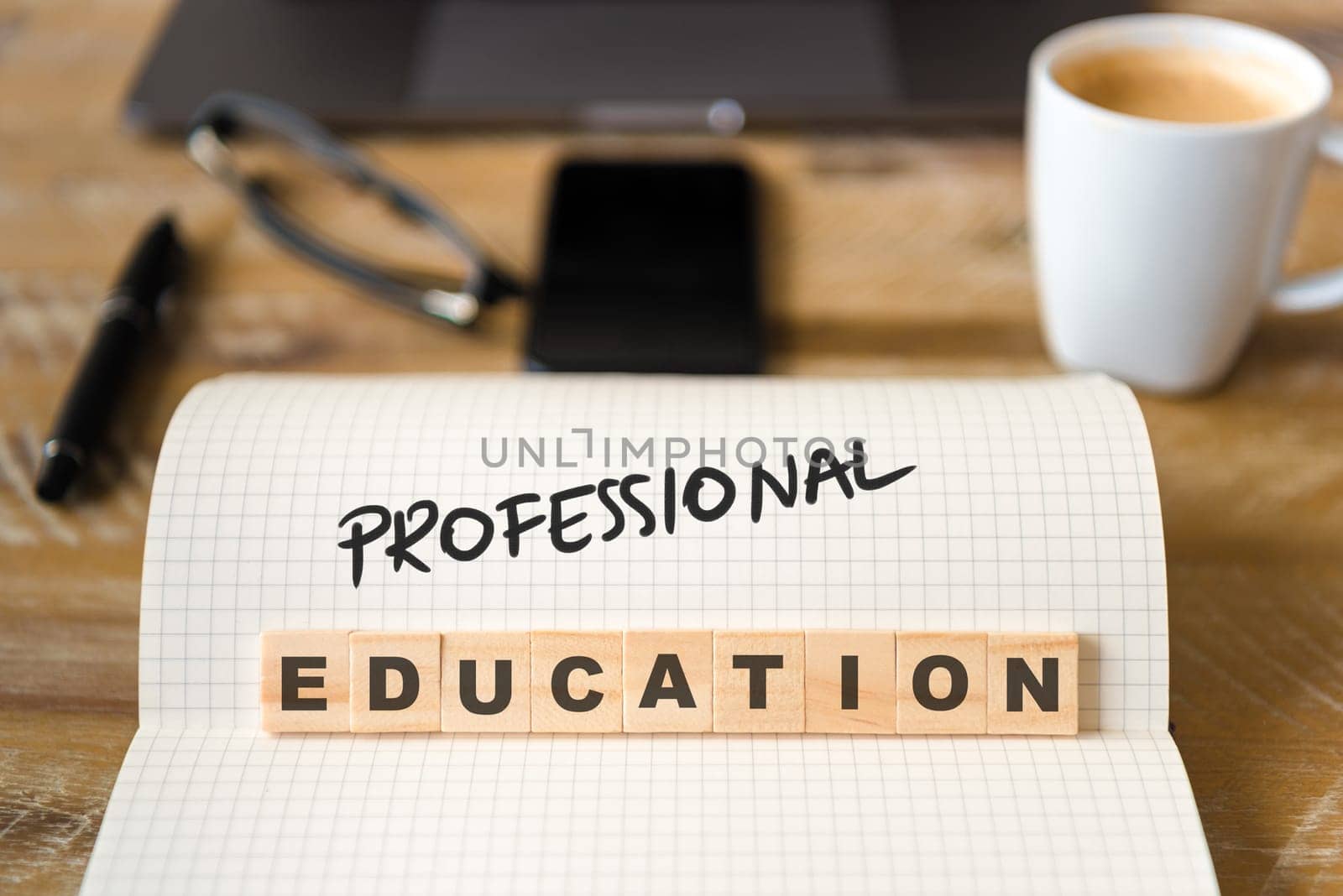 Closeup on notebook over vintage desk surface, front focus on wooden blocks with letters making Professional Education text. Business concept image with office tools and coffee cup in background