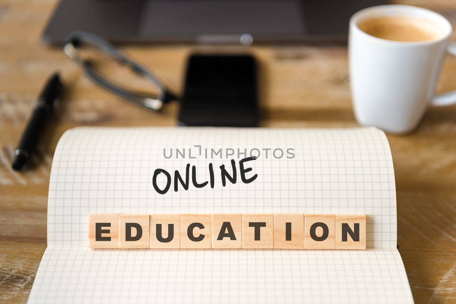 Closeup on notebook over vintage desk surface, front focus on wooden blocks with letters making Online Education text. Business concept image with office tools and coffee cup in background