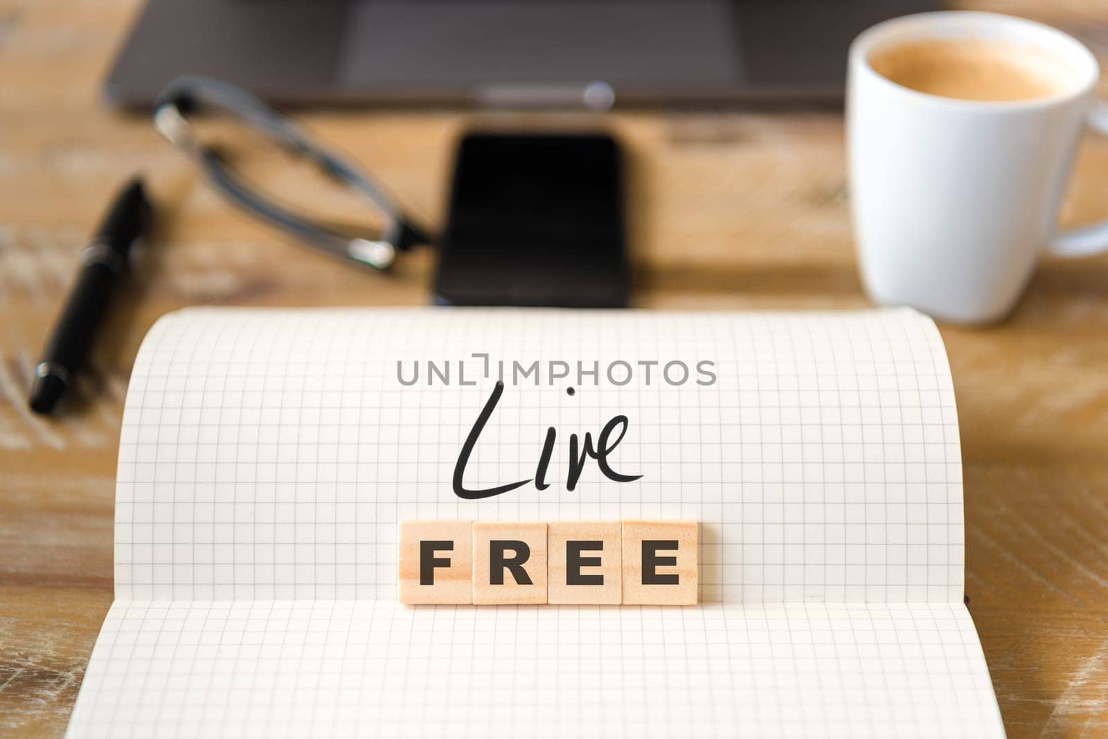 Closeup on notebook over vintage desk surface, front focus on wooden blocks with letters making Live Free text. Business concept image with office tools and coffee cup in background