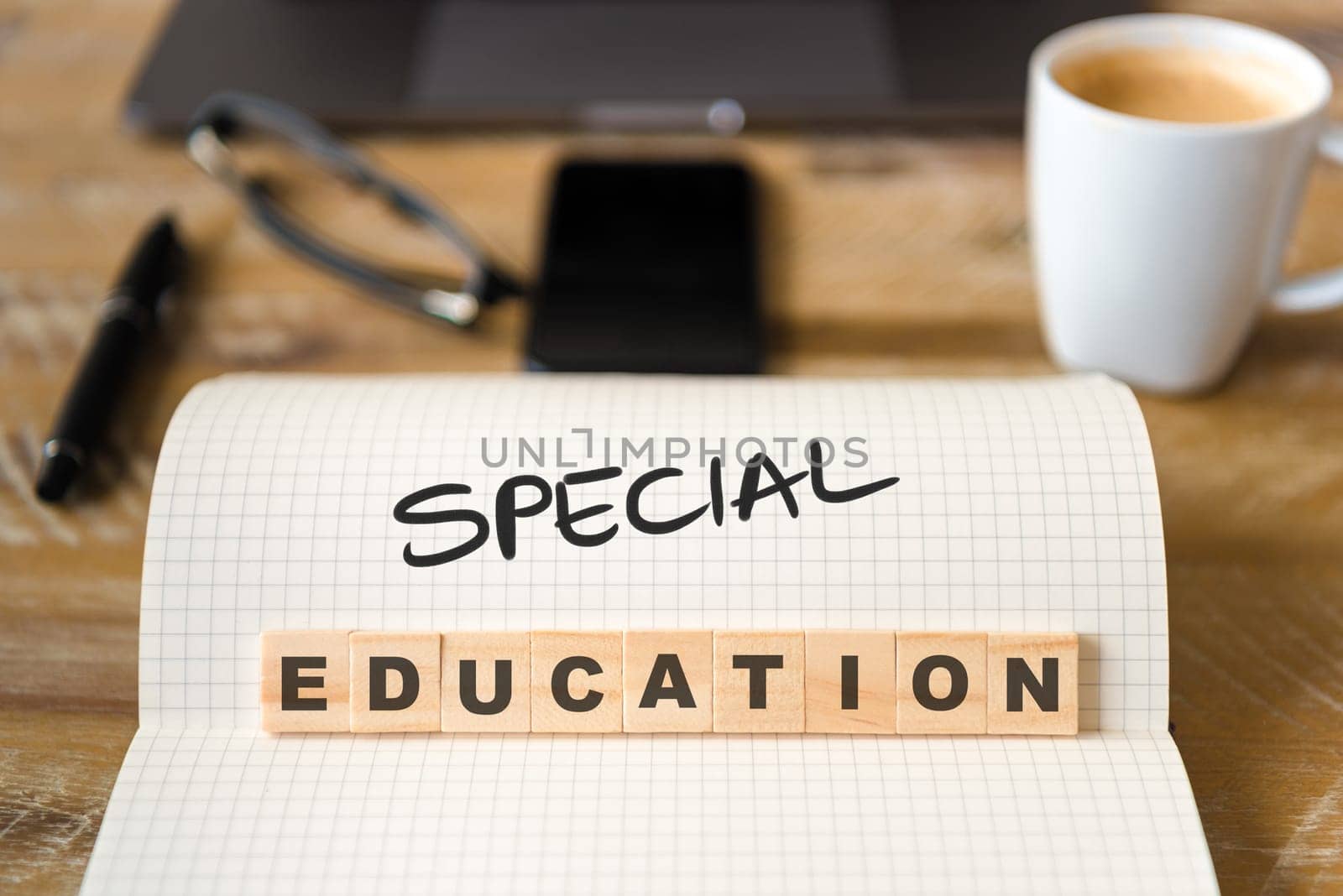 Closeup on notebook over vintage desk surface, front focus on wooden blocks with letters making Special Education text. Business concept image with office tools and coffee cup in background