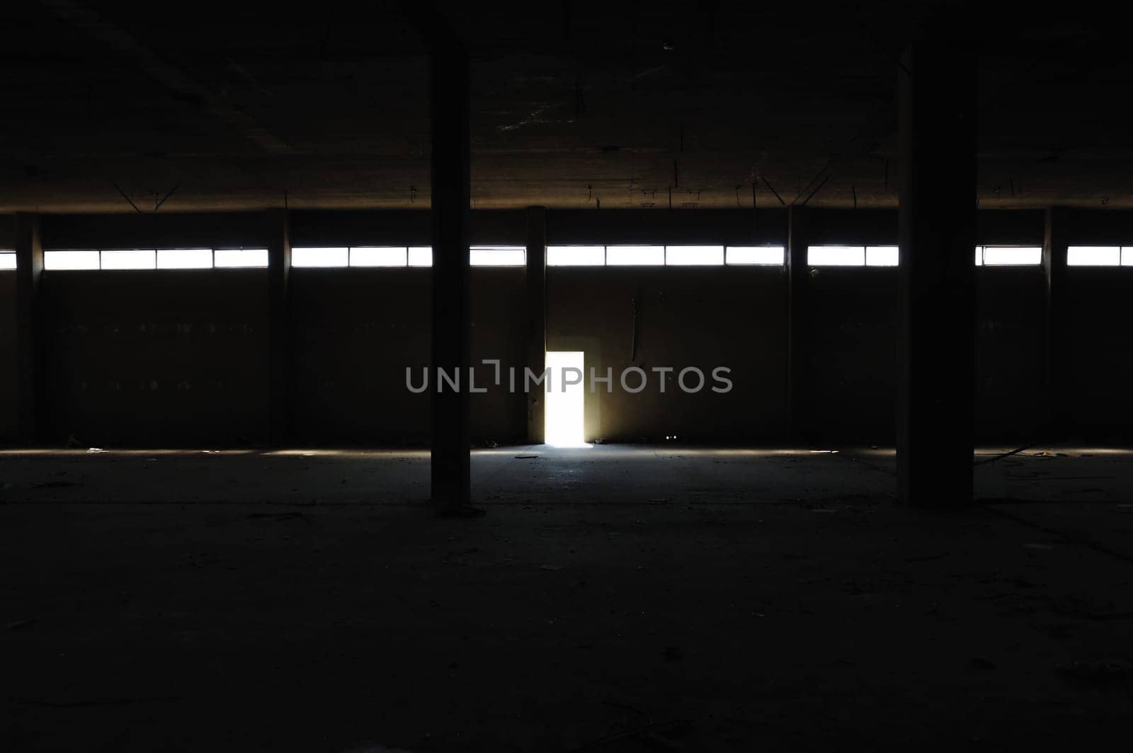 Dark interior in abandoned factory concrete pillars and light by the exit door. Industrial ruin.