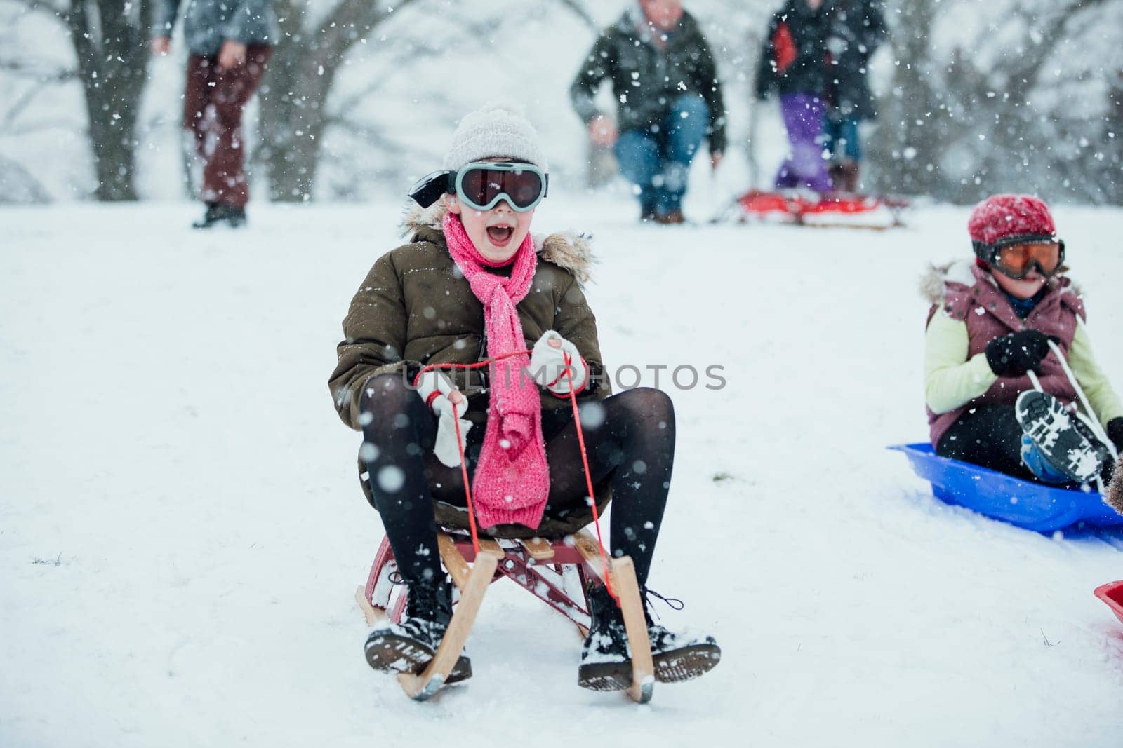 Little girl is going down a big snowy hill on a sleigh while wearing ski goggles.