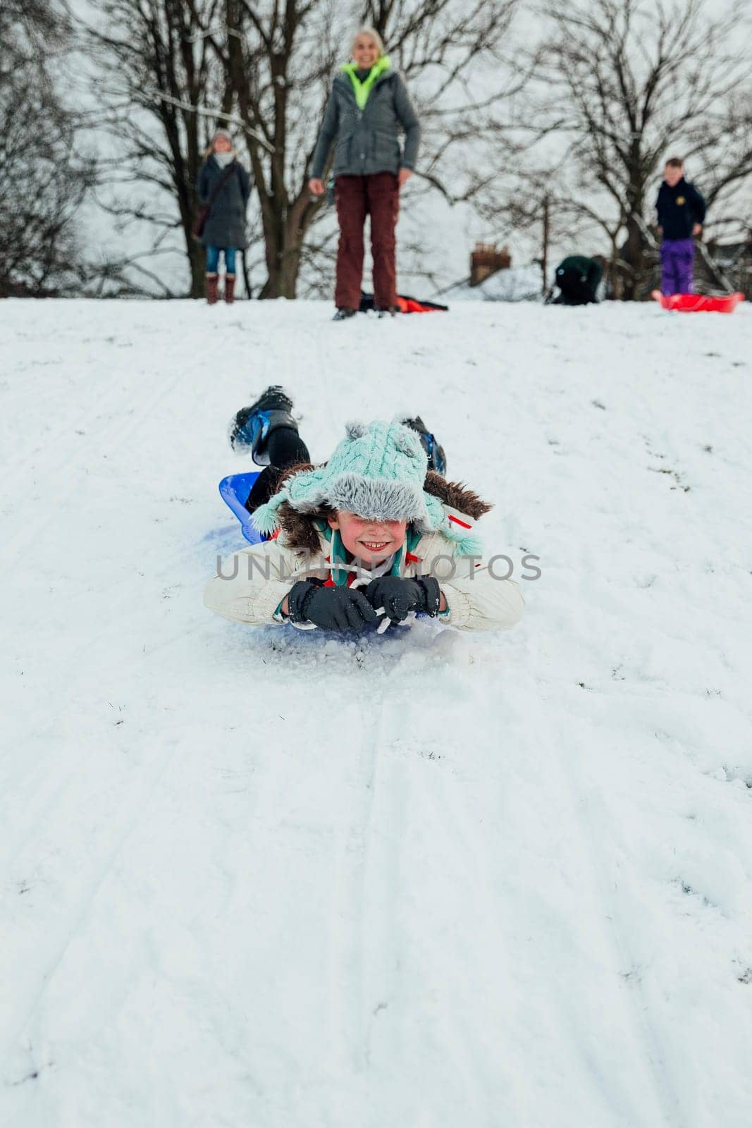 Little girl is lying on her belly on a sled, zooming down a big hill in the snow.