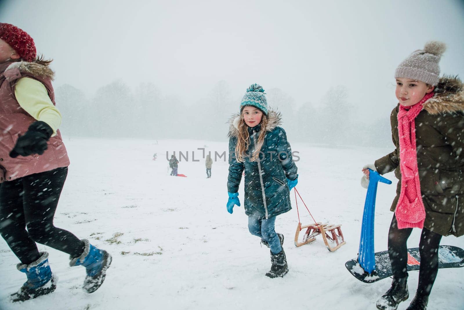 Little girls are walking up a hill with sleds in a snow blizzard to have a race.